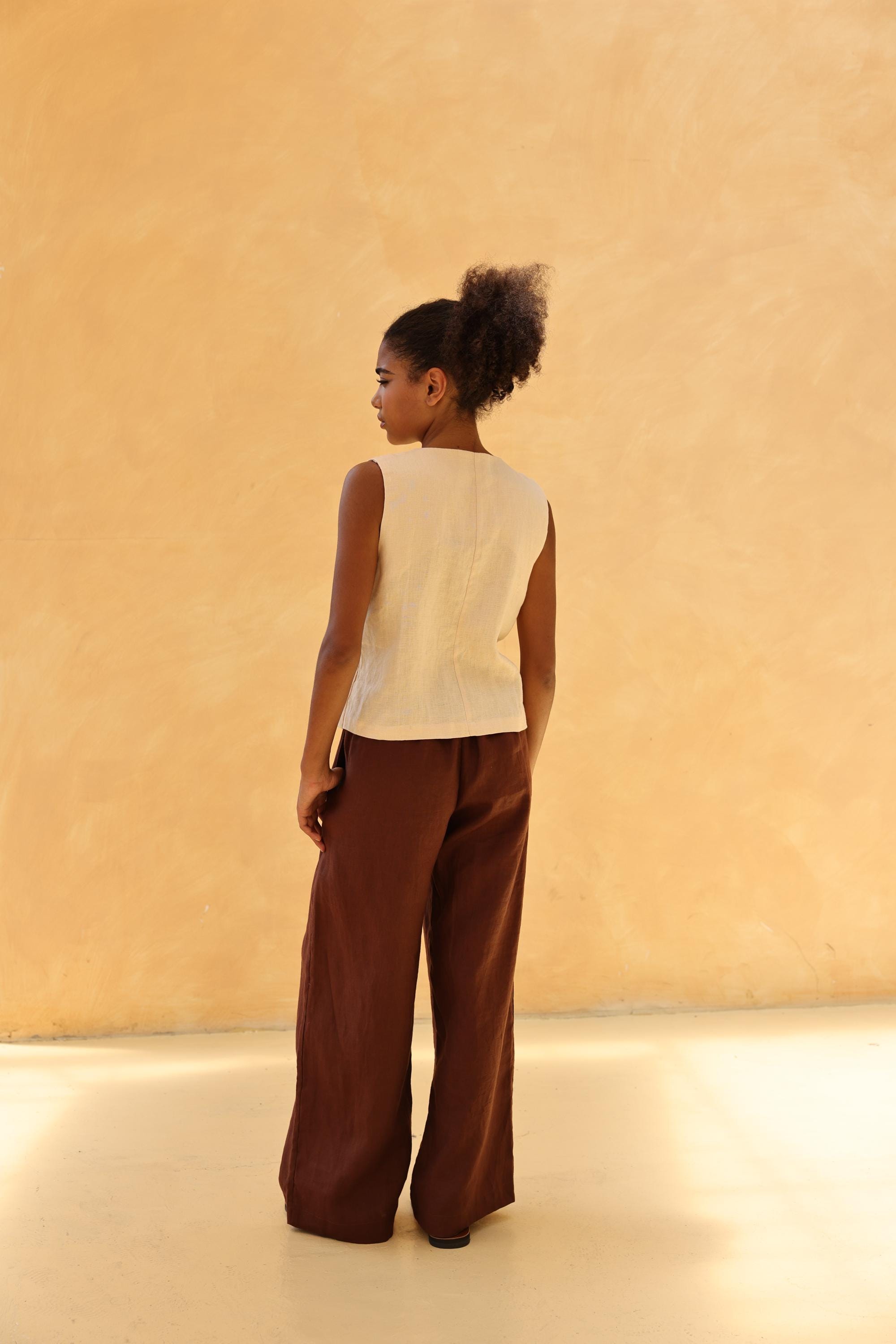 Woman wearing a beige sleeveless top and brown wide-leg pants against a beige wall.