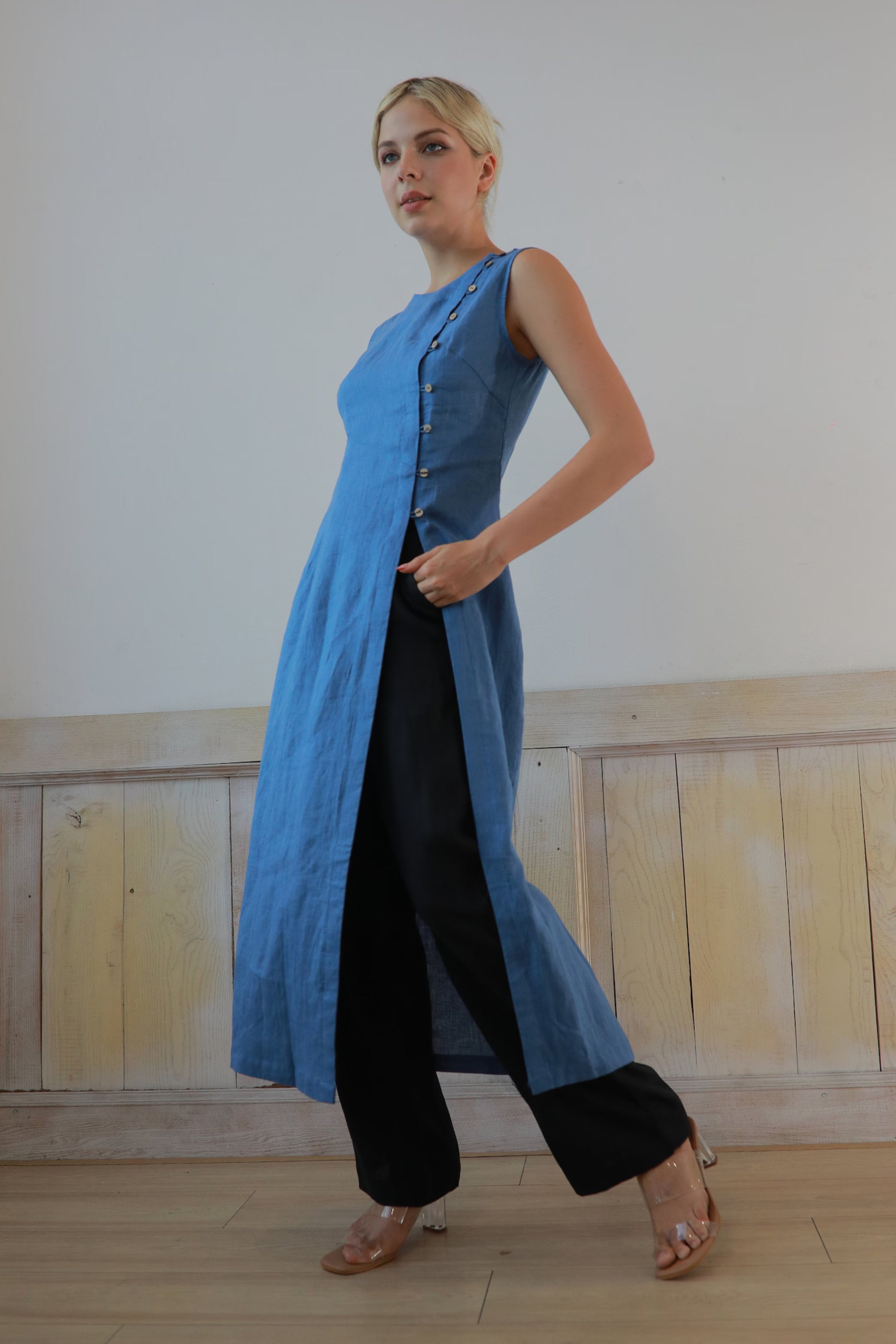 Woman wearing a blue dress with black pants against a plain wall.