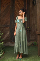 Woman wearing a green dress with floral embroidery standing in front of a wooden door.