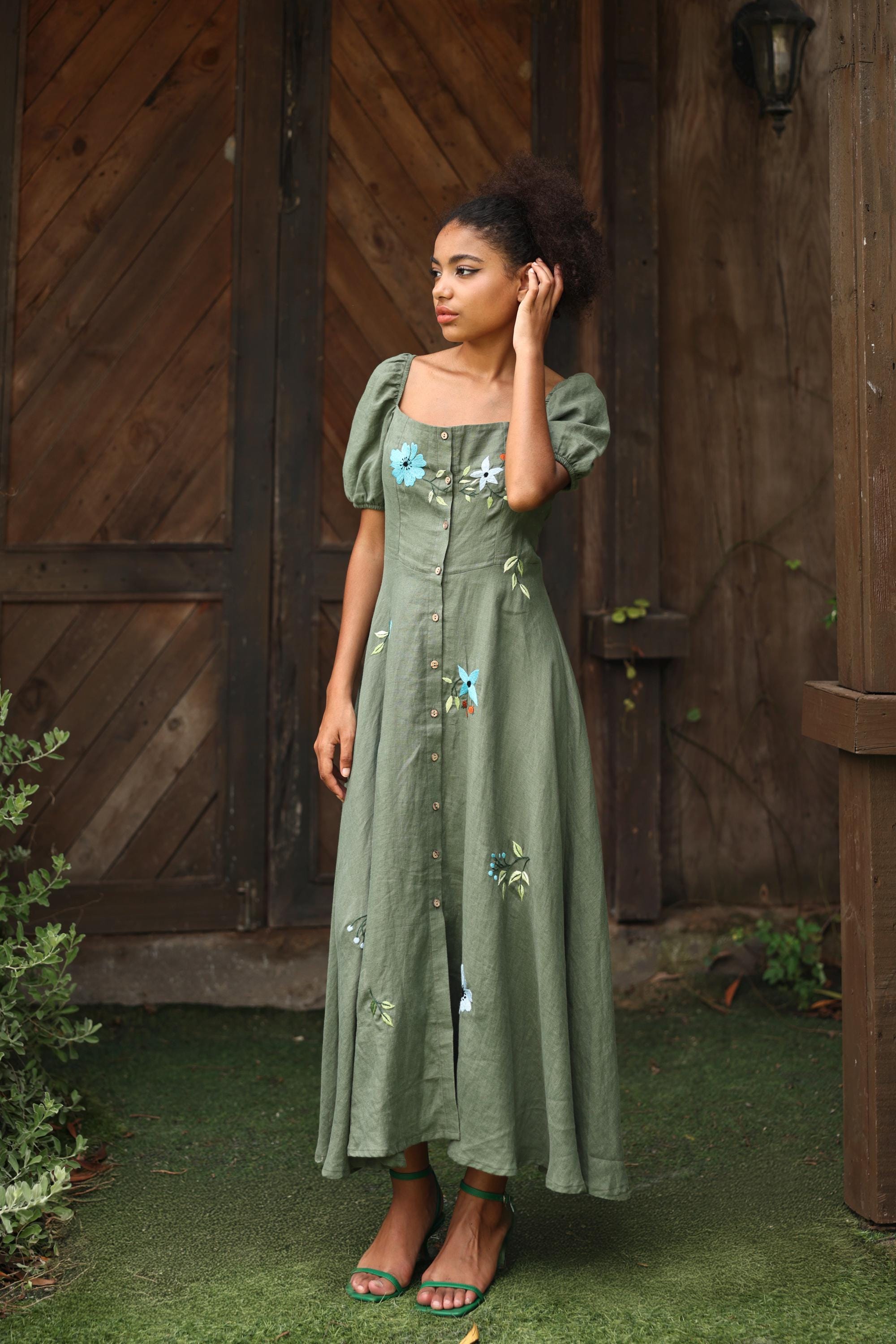 Woman wearing a green dress with floral embroidery standing in front of a wooden door.