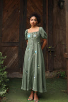 Woman in a green dress with floral embellishments standing in front of a wooden door.