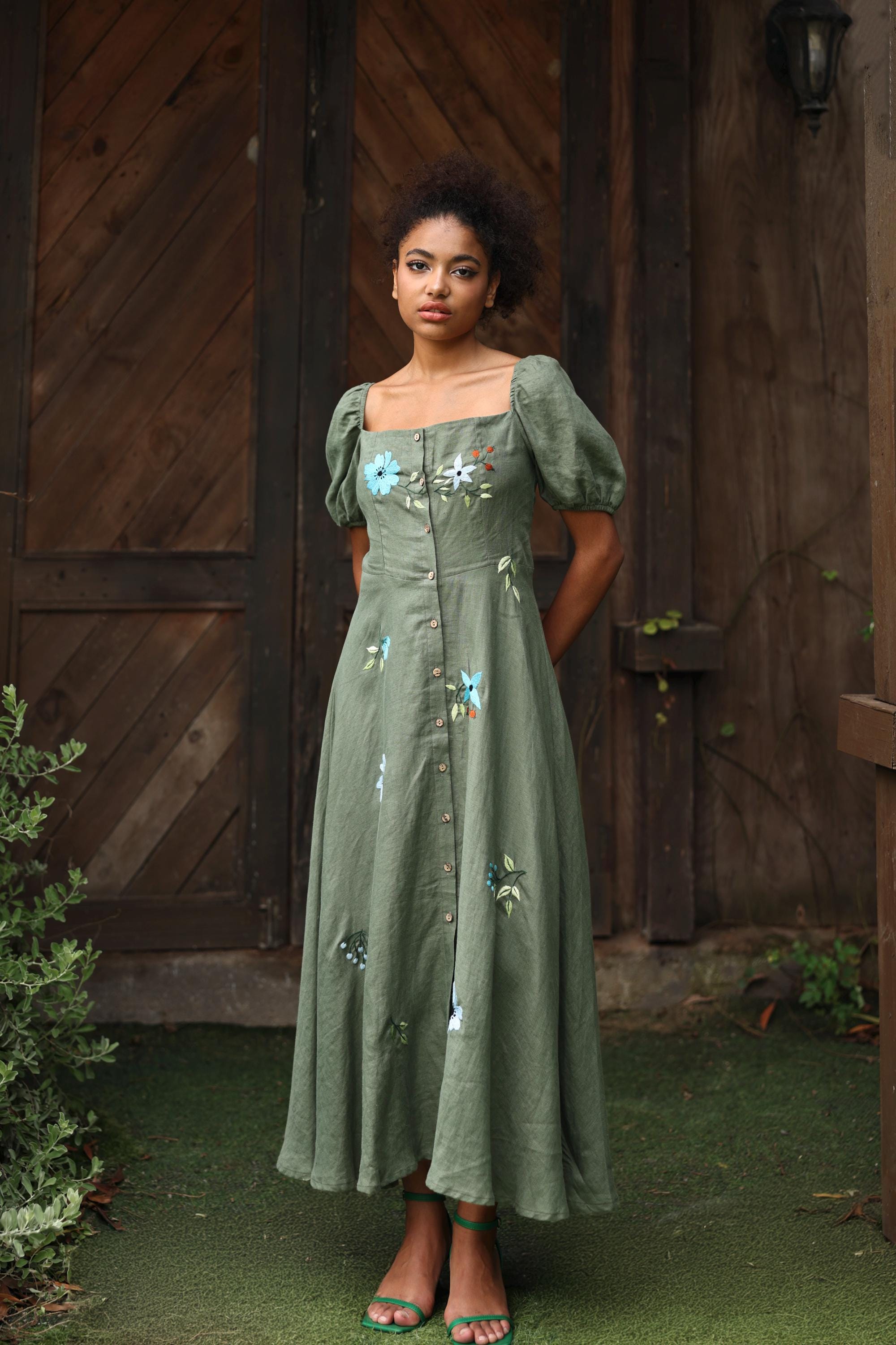 Woman in a green dress with floral embellishments standing in front of a wooden door.