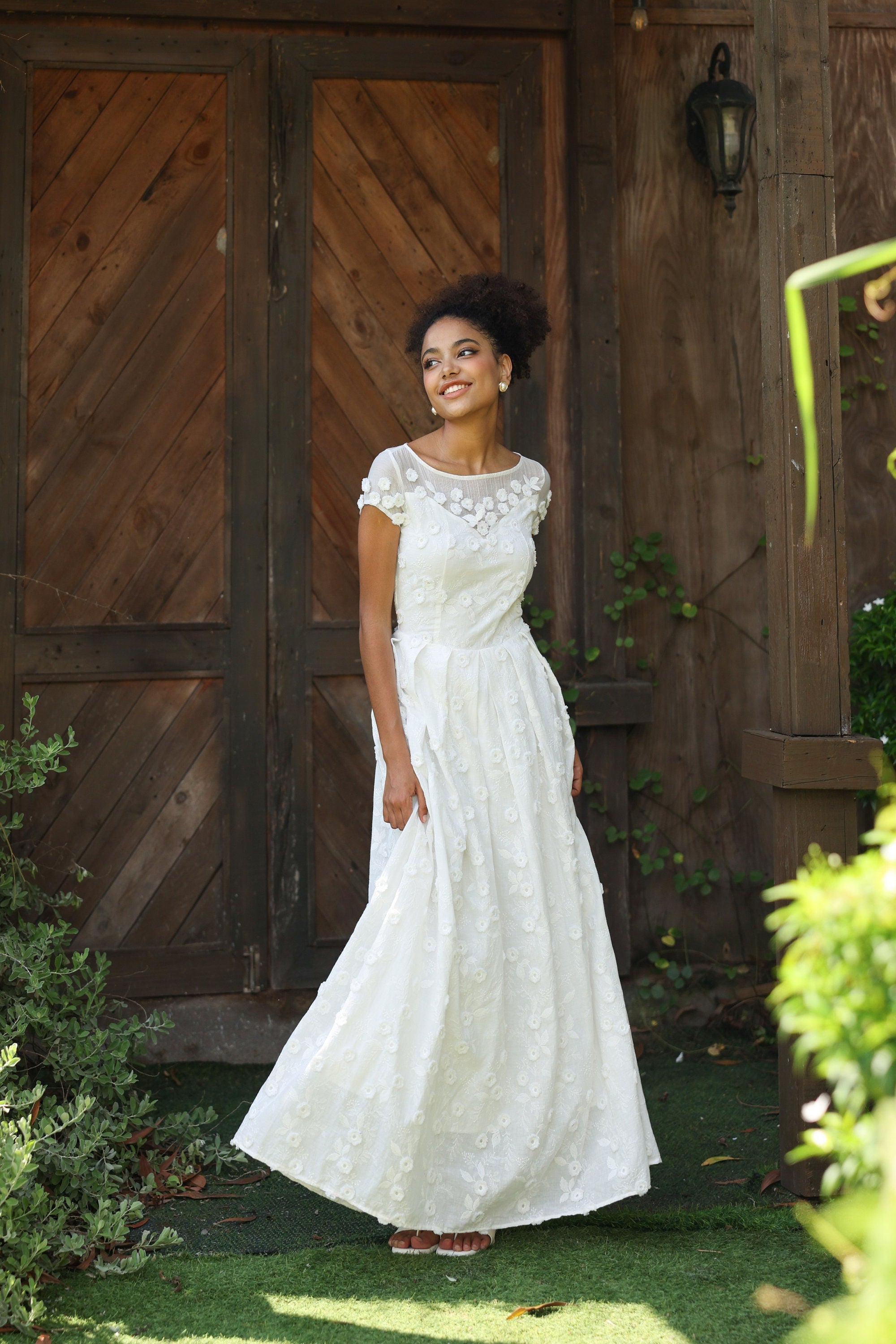 Woman in a white lace dress standing in front of wooden doors with greenery around.