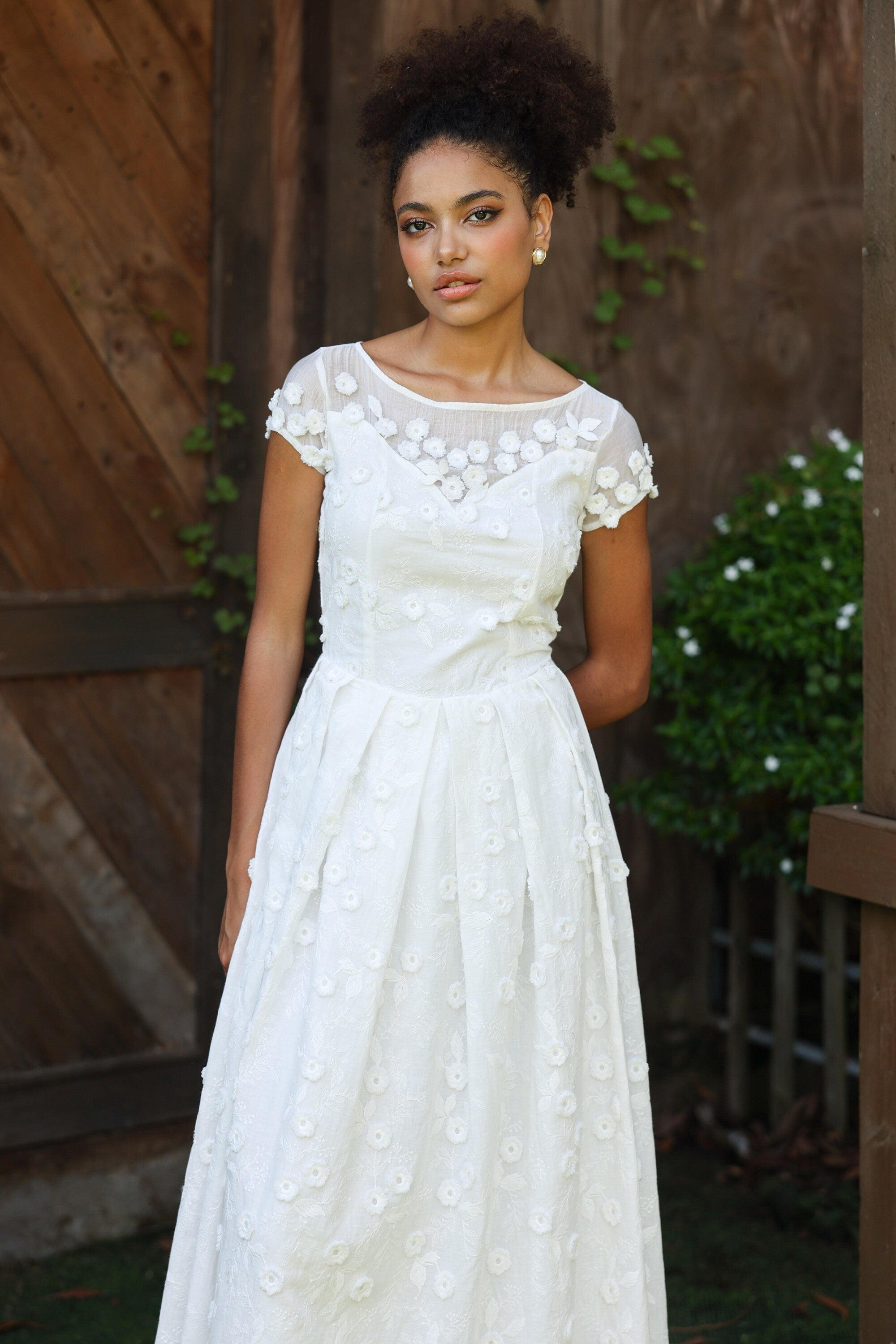 Woman wearing a white lace dress standing outdoors with wooden and greenery background