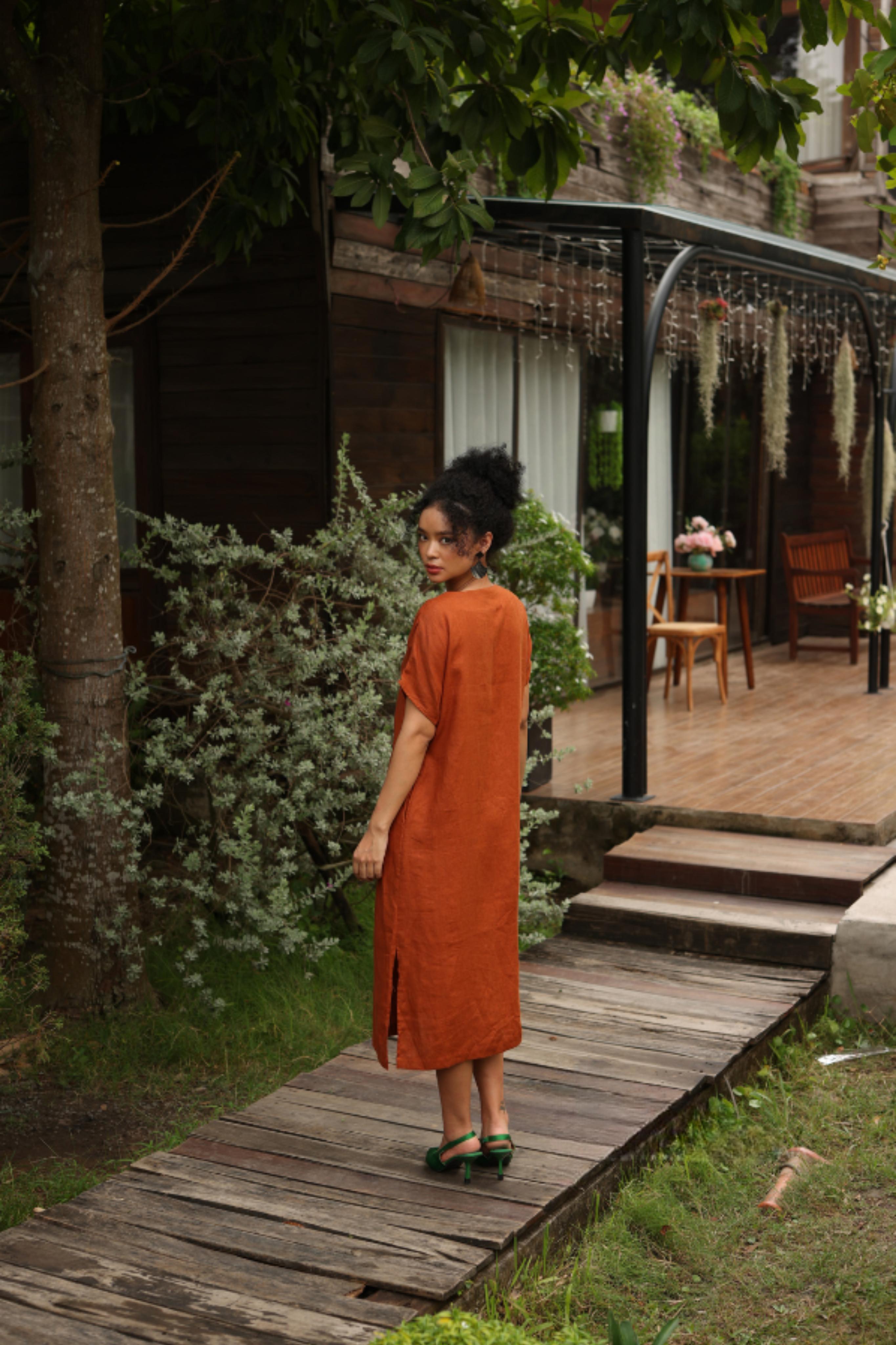 Woman in a dress standing on a wooden path in a garden setting.