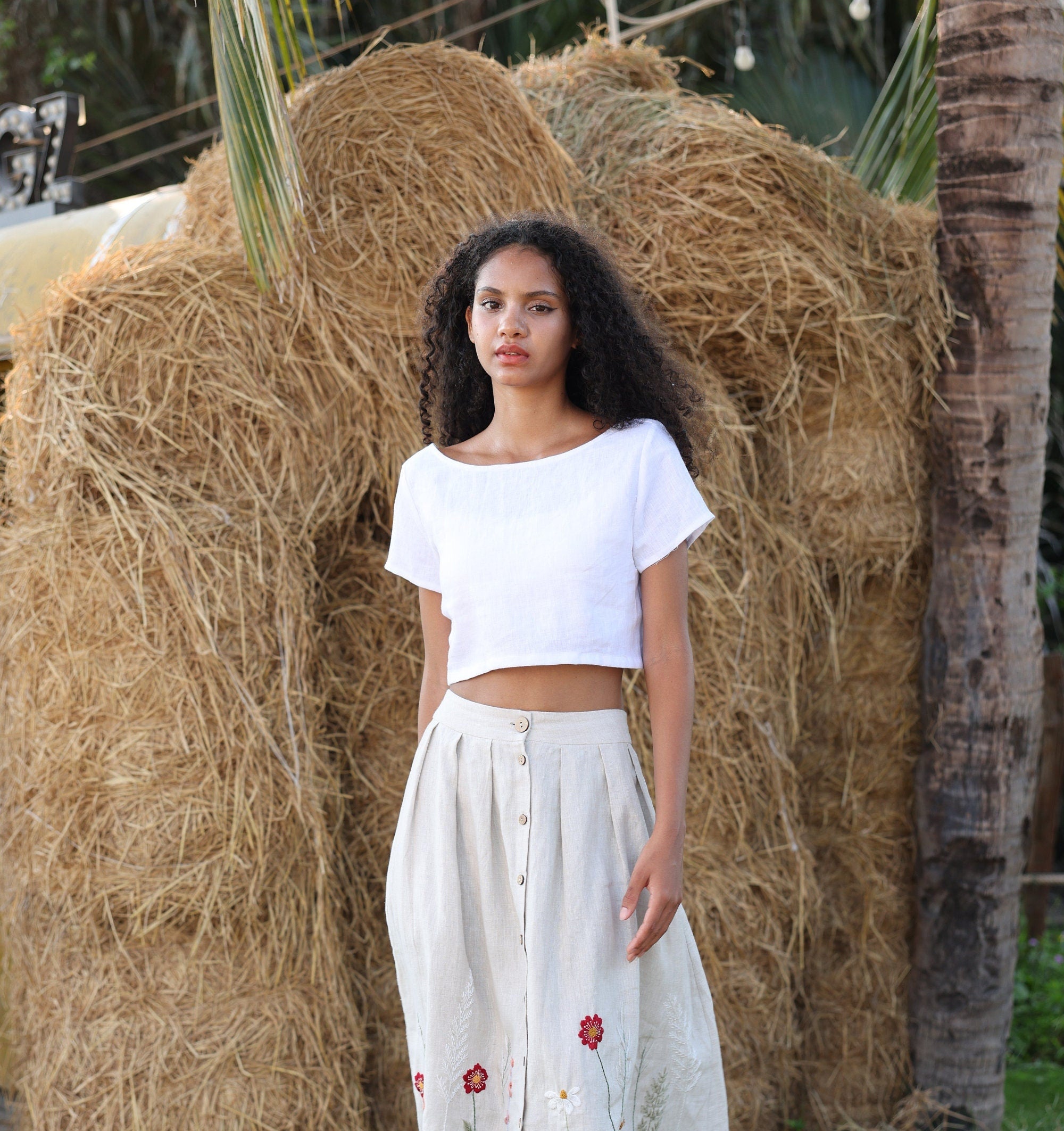 Woman in a white top and light-colored skirt standing in front of hay bales.