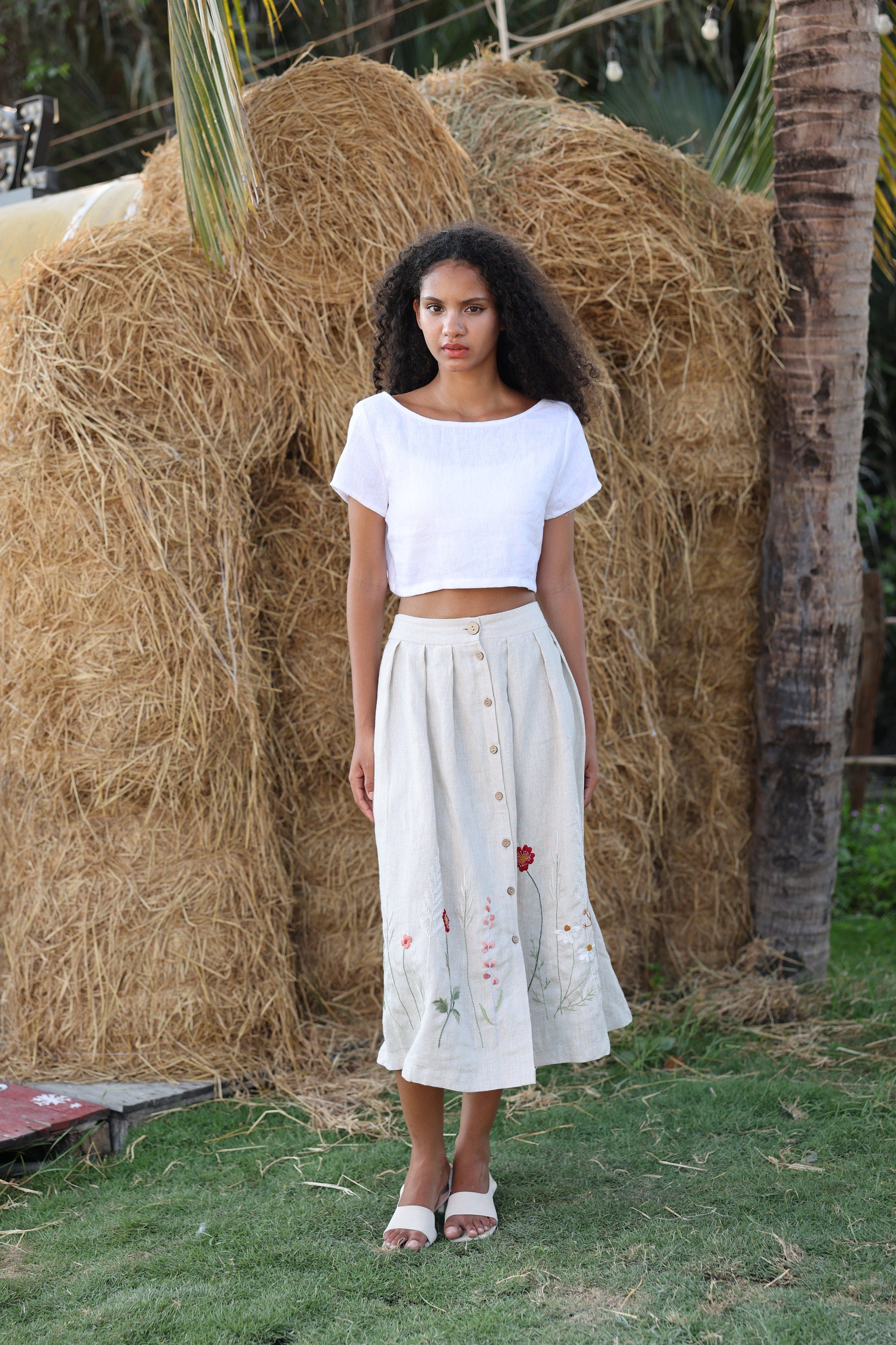 Woman in a white top standing in front of hay bales with palm trees in the background