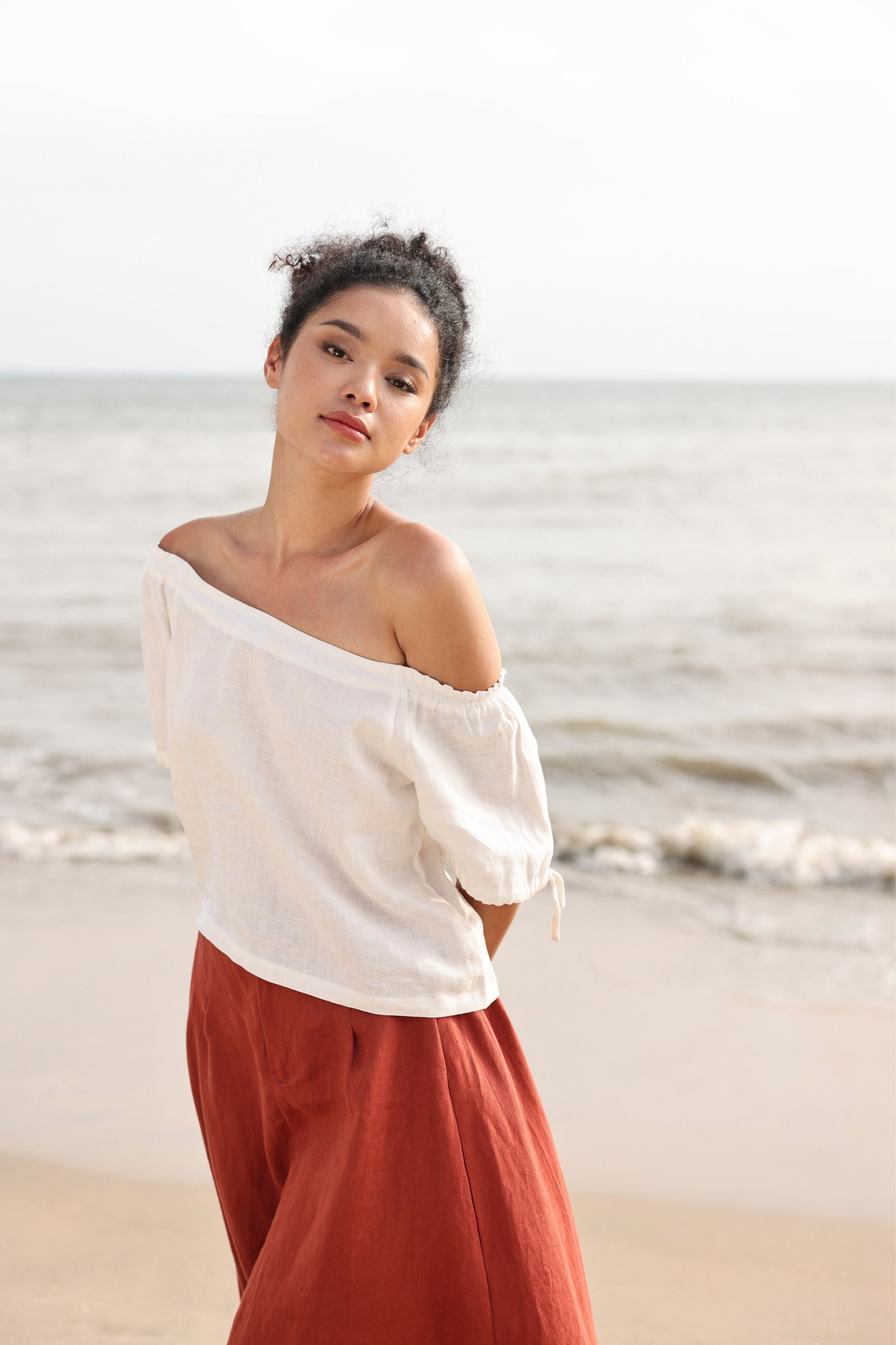 Woman wearing a white off-shoulder top and red skirt standing on a beach.