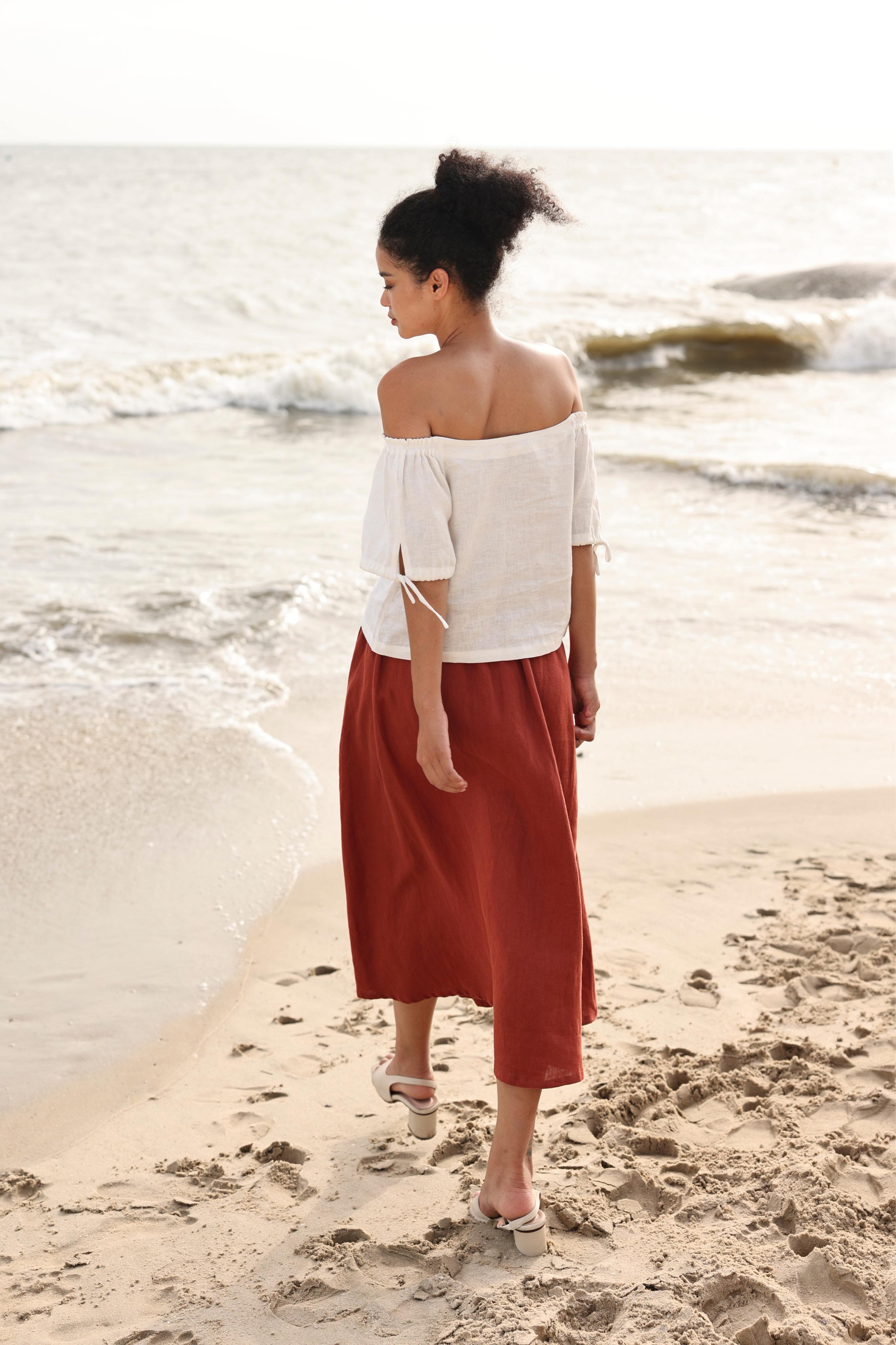 Woman walking on a beach wearing a white off-shoulder top and red skirt.
