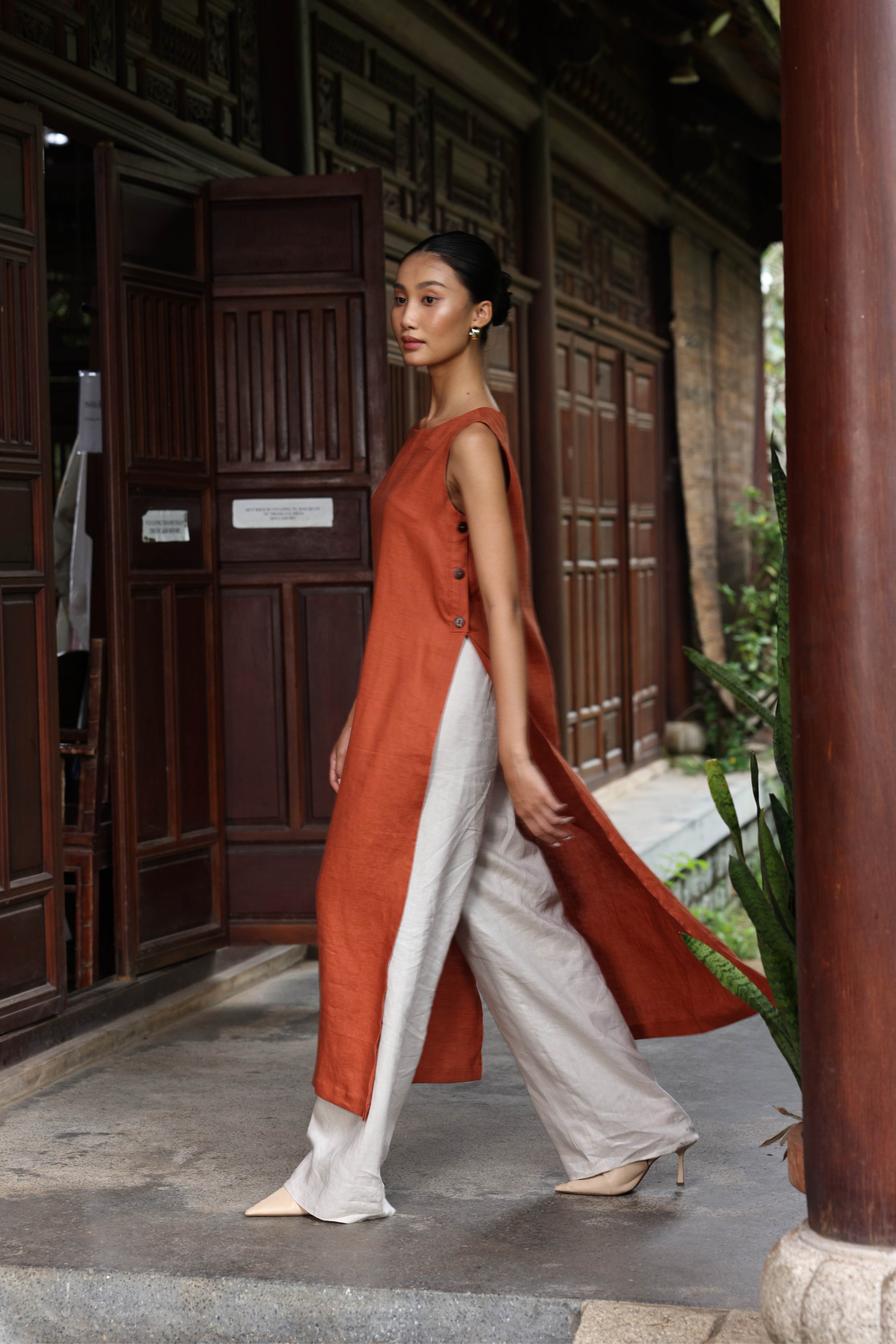 Woman in a rust-colored dress and white pants standing in front of traditional wooden doors.