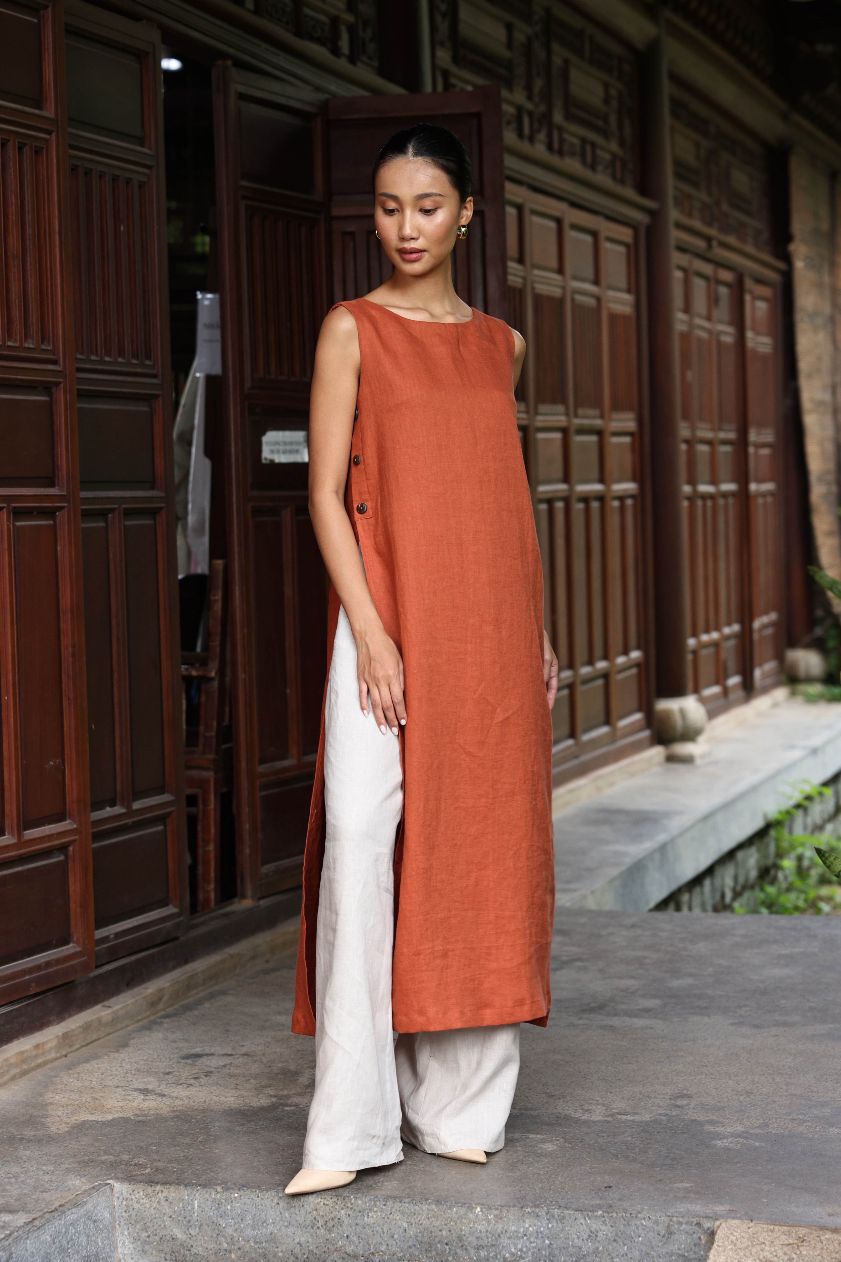 Woman wearing a long rust-colored dress with white pants standing in front of a traditional wooden building.