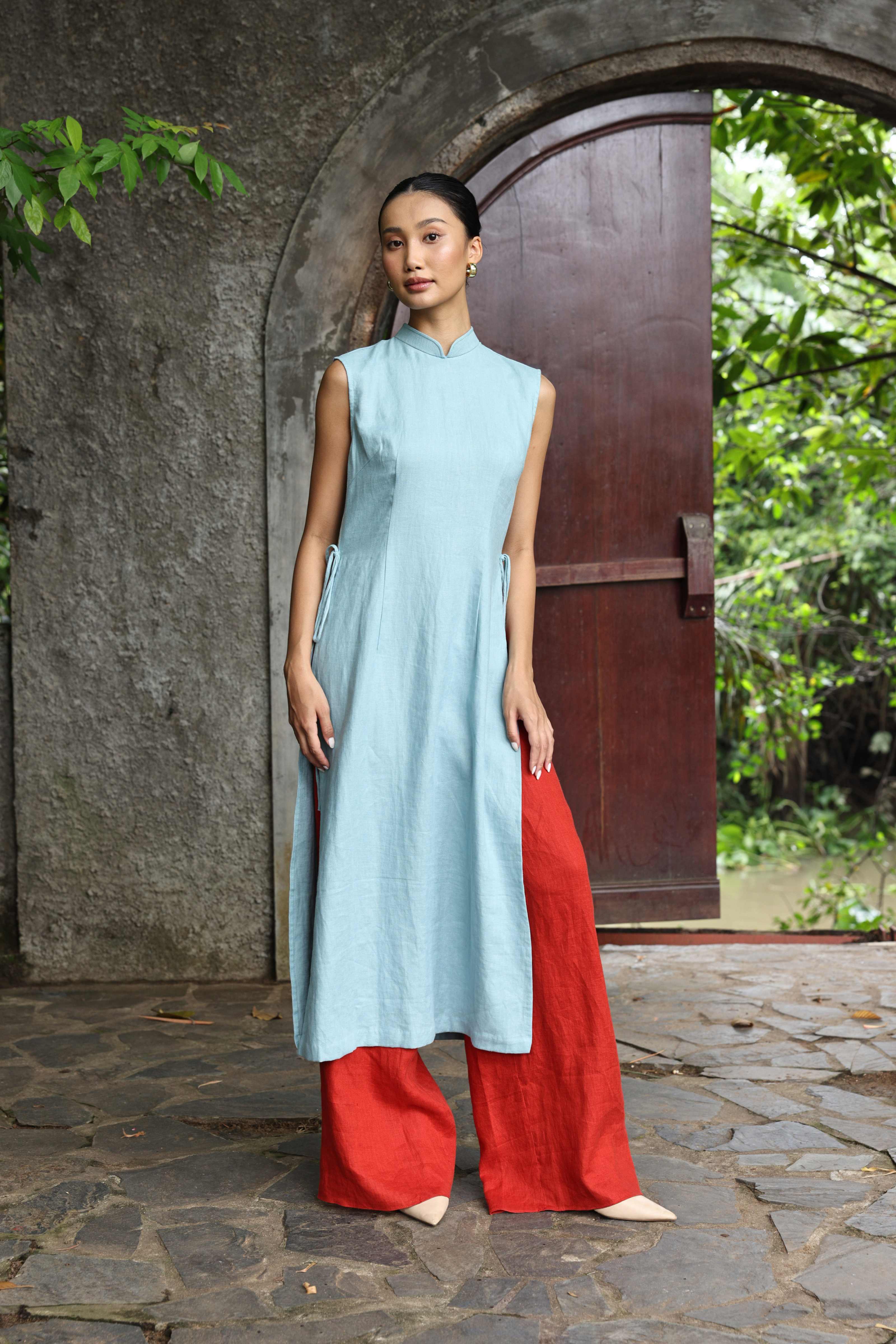 Woman wearing a light blue sleeveless top and red pants standing in front of a stone archway with greenery.