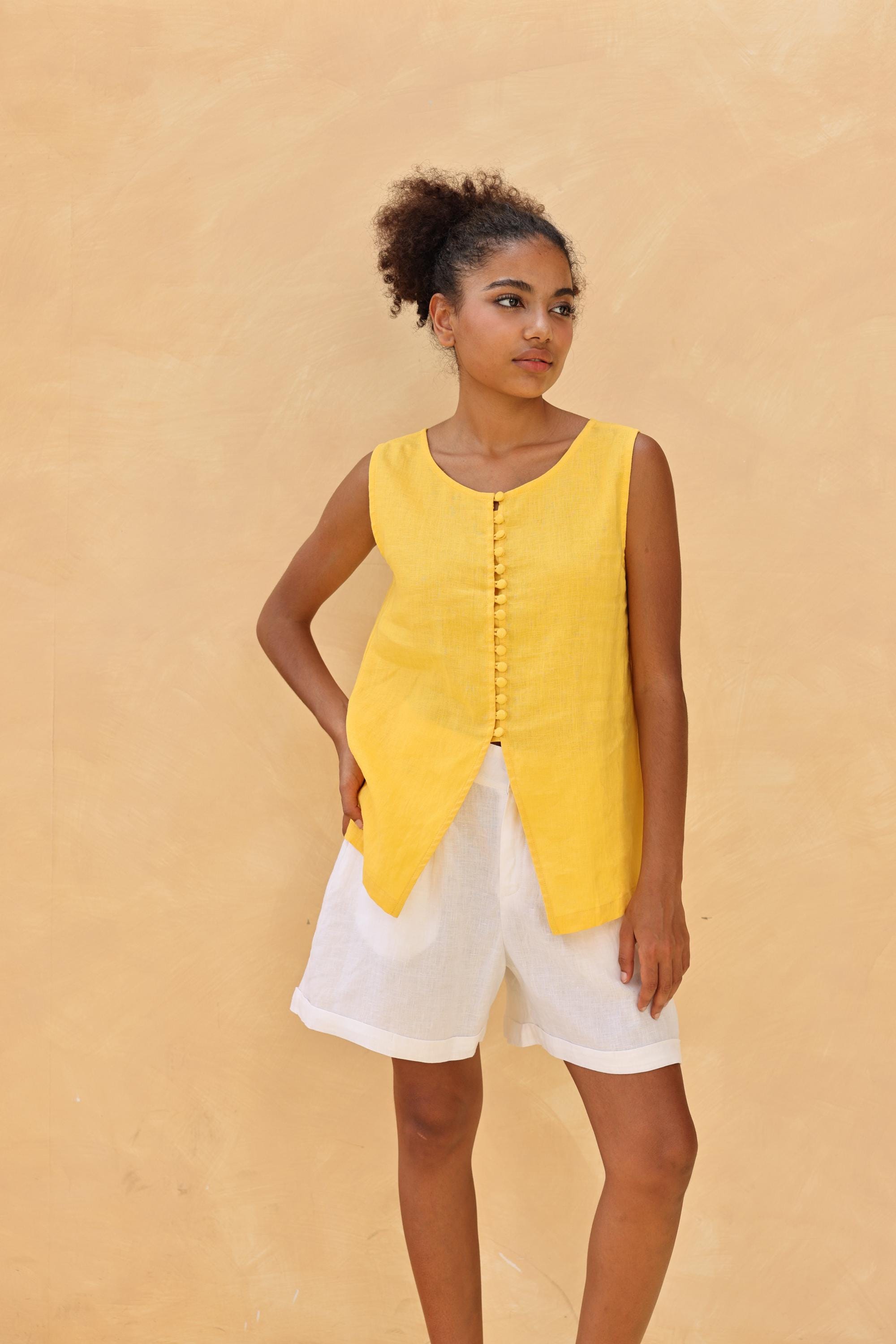 Woman wearing a yellow sleeveless top and white shorts against a beige background