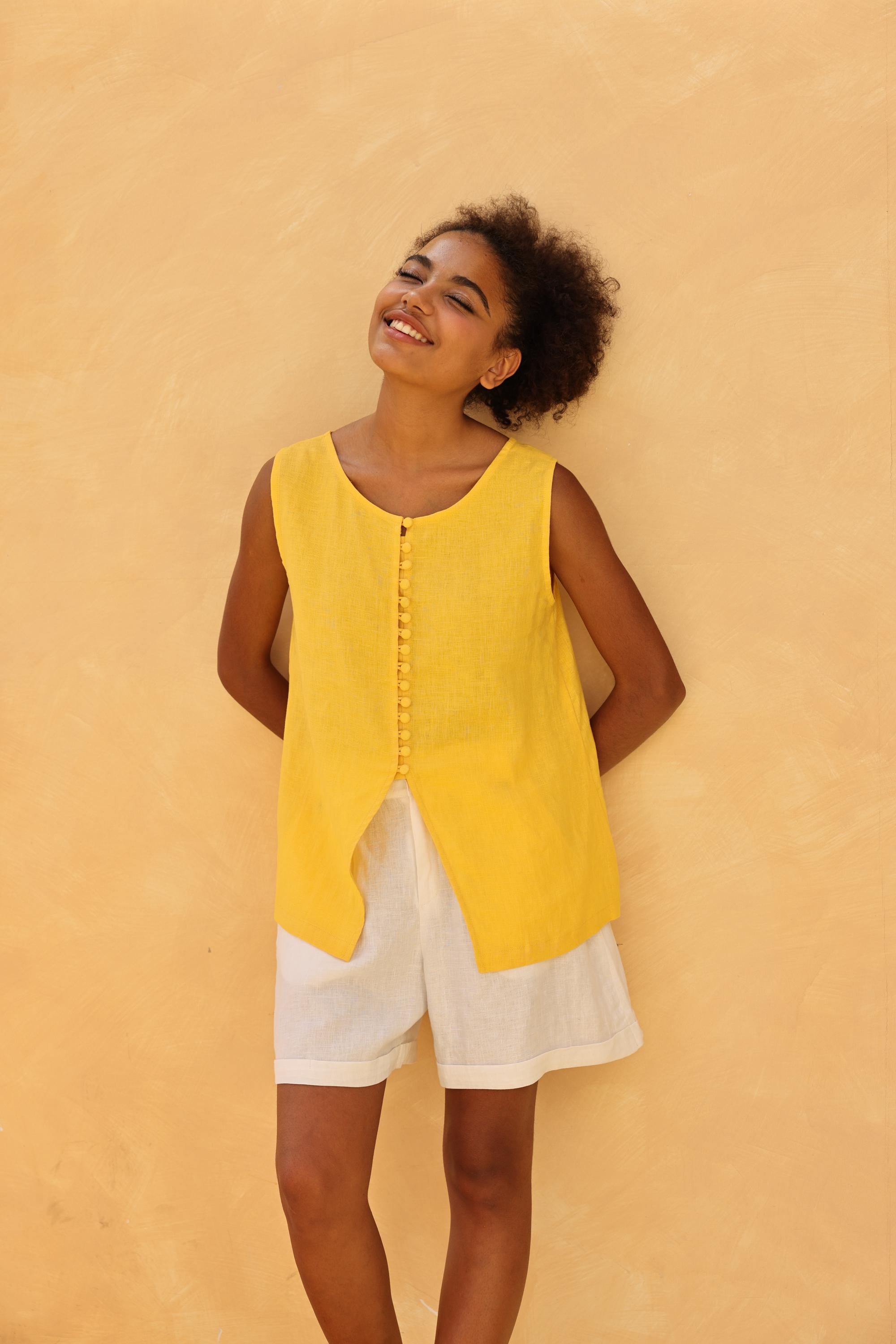 Woman wearing a yellow sleeveless top and white shorts against a beige wall
