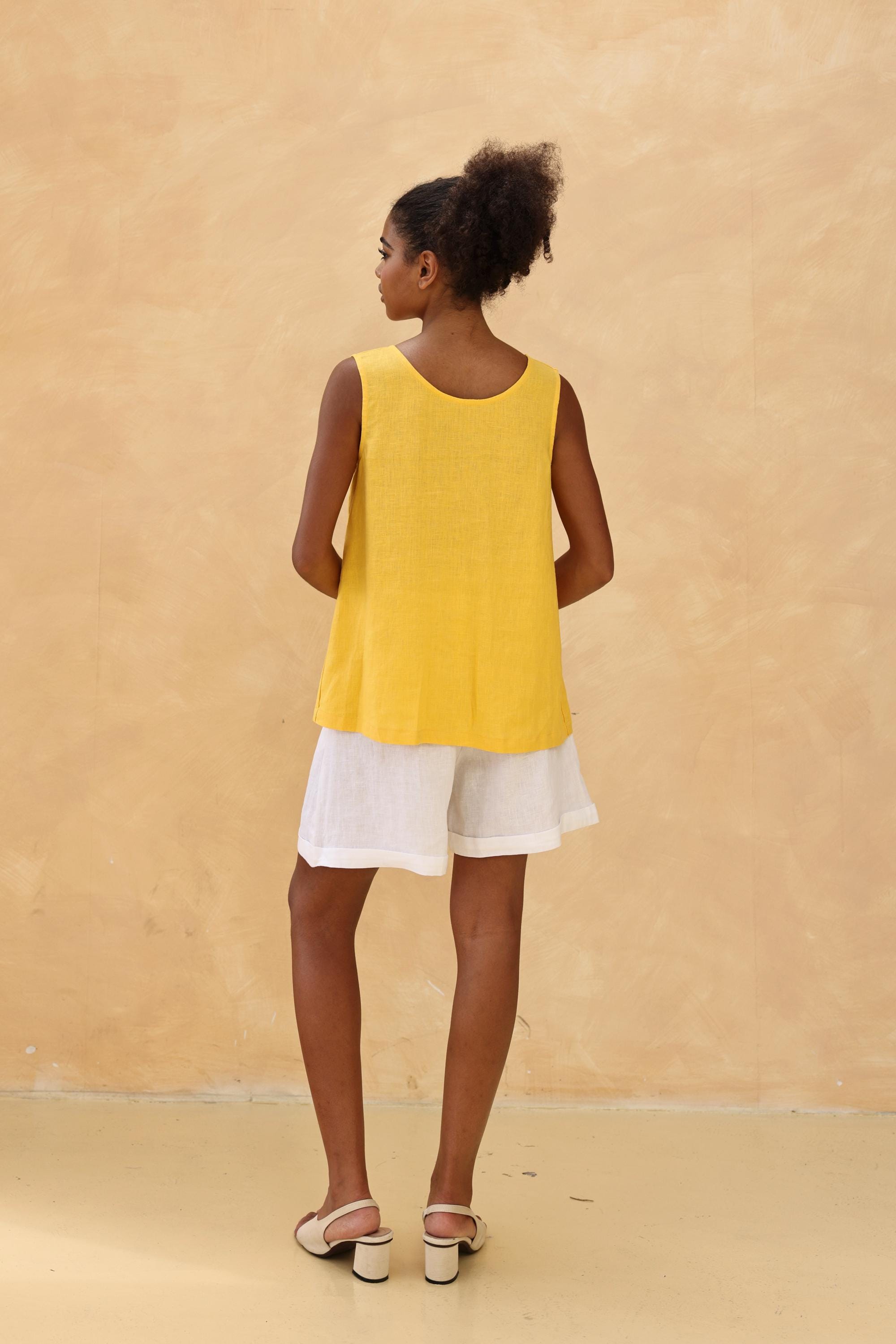 Woman wearing a yellow sleeveless top and white shorts against a beige background