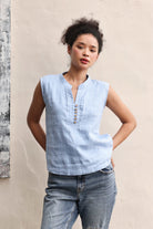 Woman wearing a light blue sleeveless shirt and jeans against a beige wall.