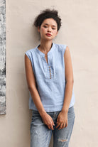 Woman wearing a light blue sleeveless top and jeans against a beige wall.