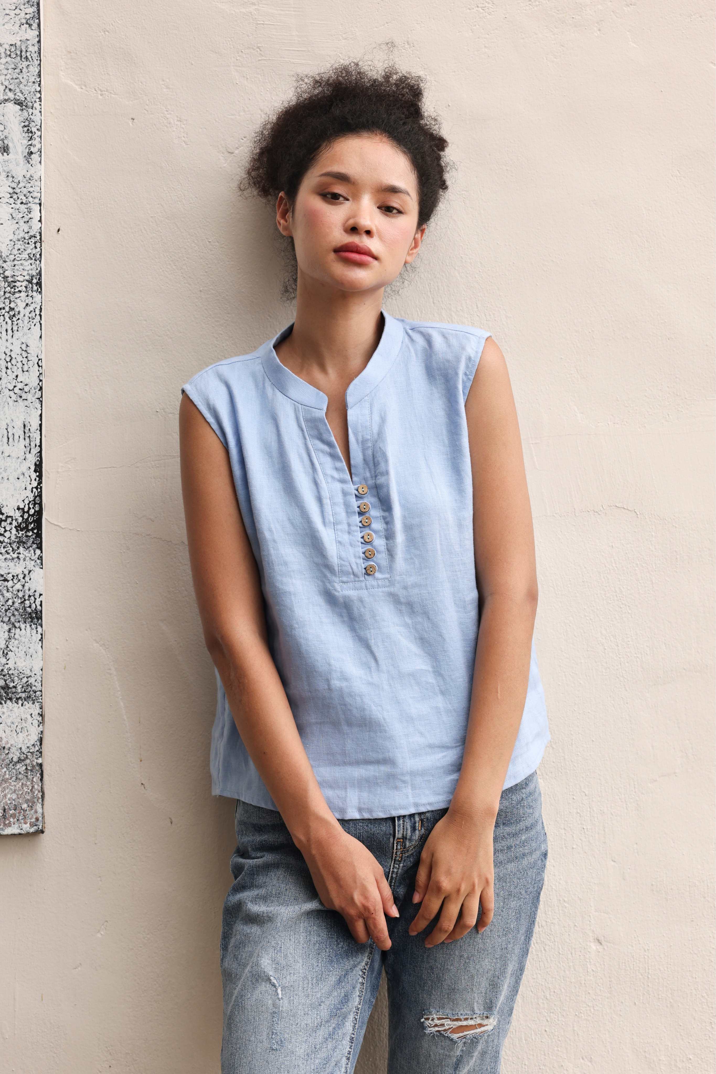 Woman wearing a light blue sleeveless top and jeans against a beige wall.