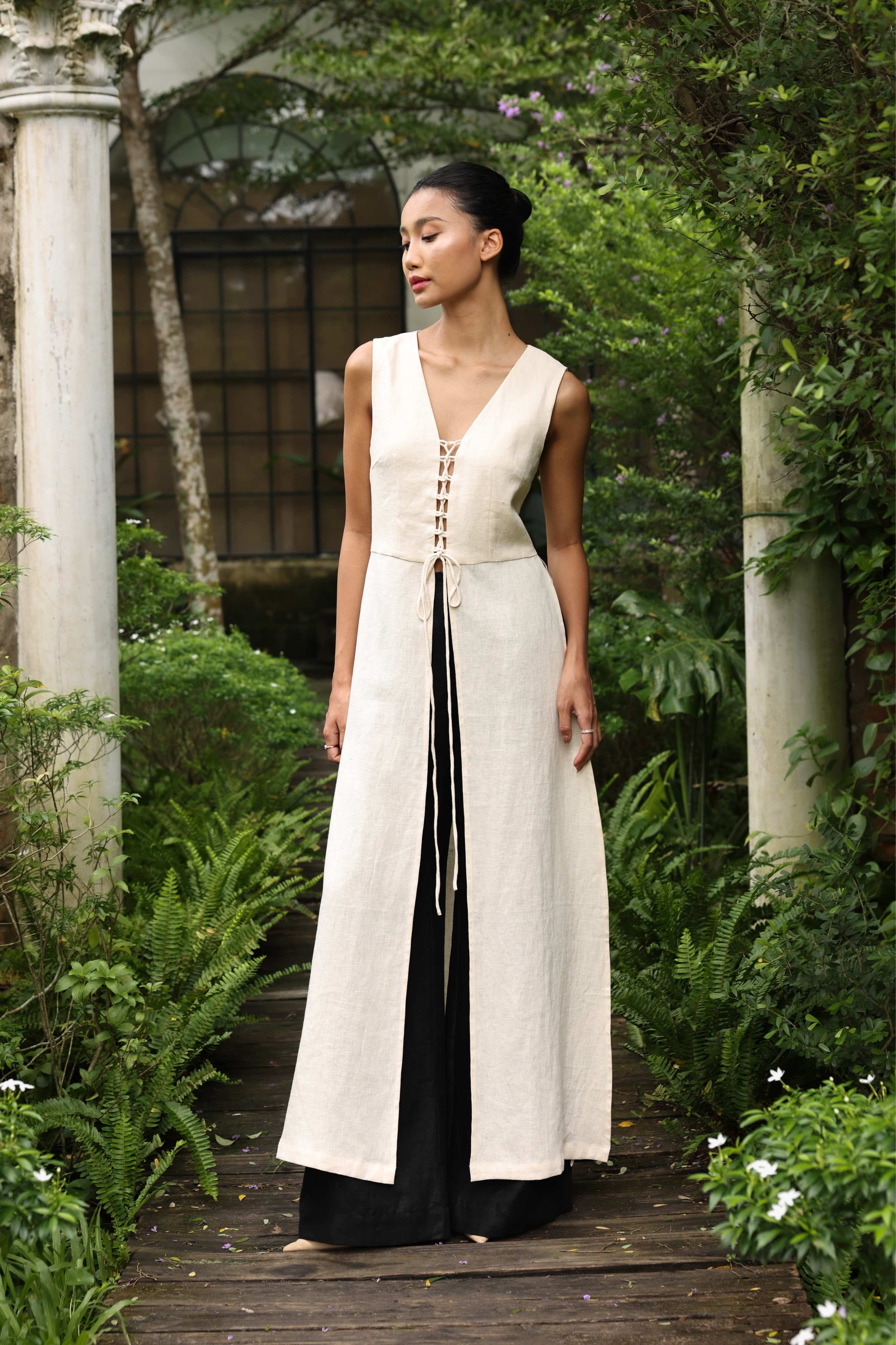 Woman in a long, light-colored dress standing in a garden setting with greenery and columns.