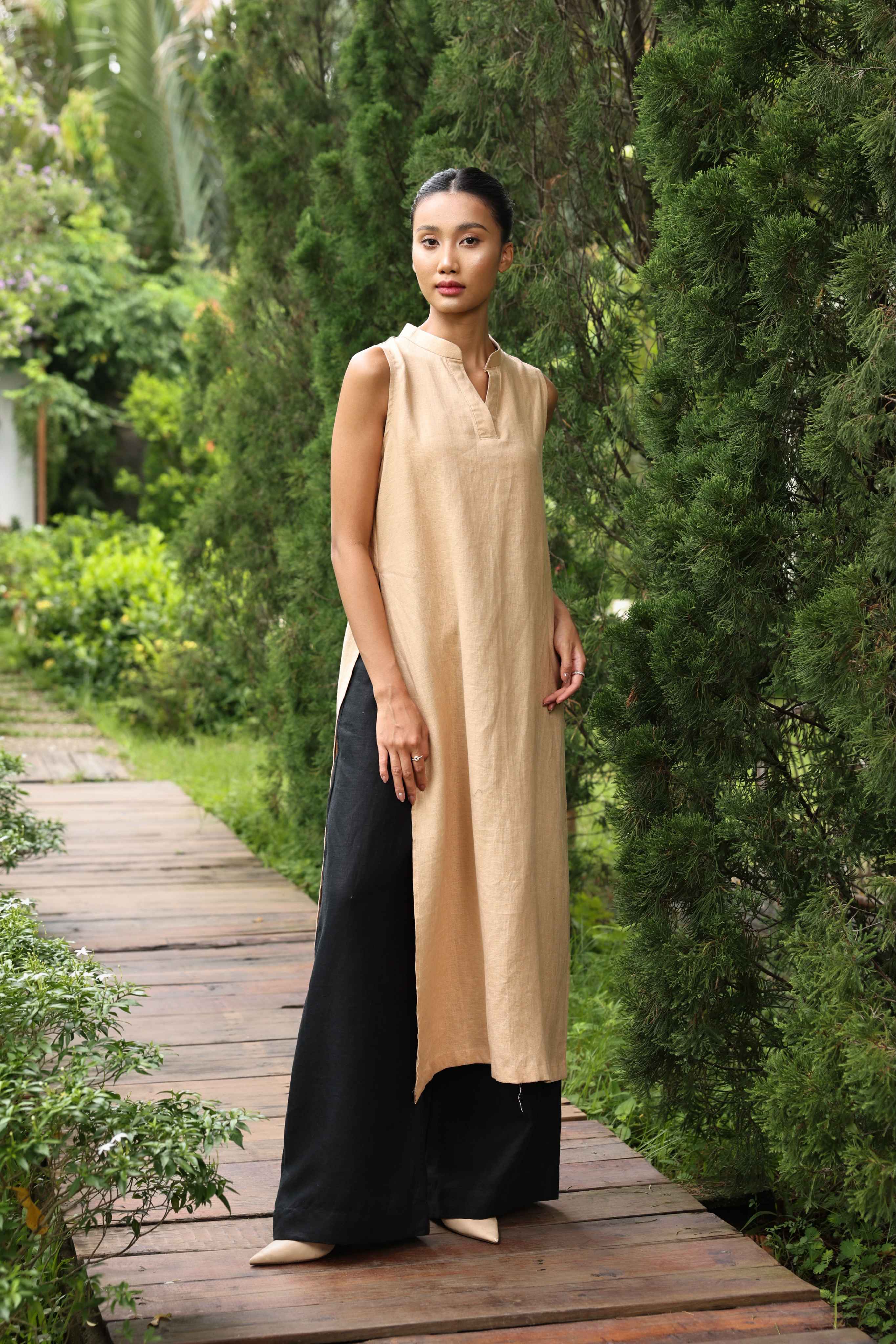 Woman wearing a beige sleeveless top and black pants standing on a wooden path in a garden.