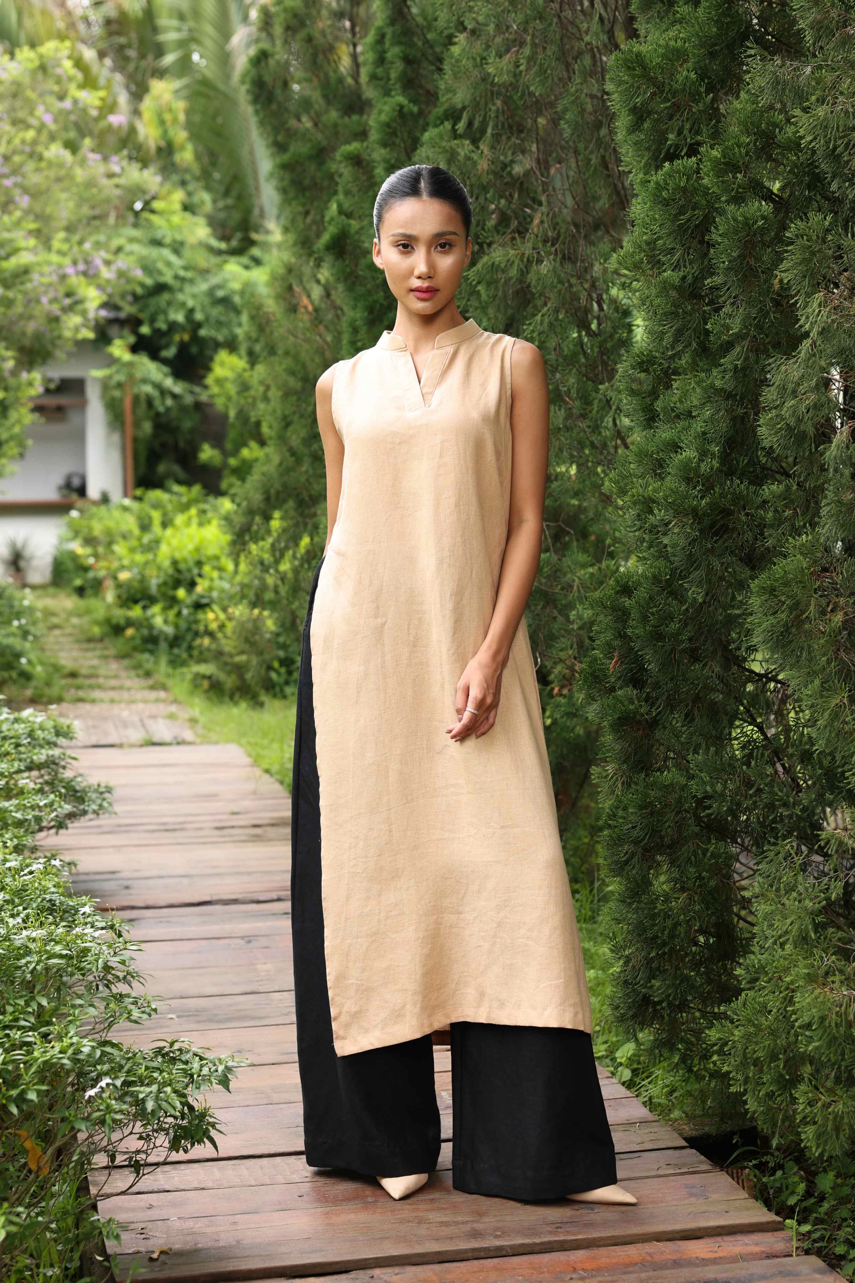 Woman in a beige sleeveless dress standing on a wooden path in a garden.