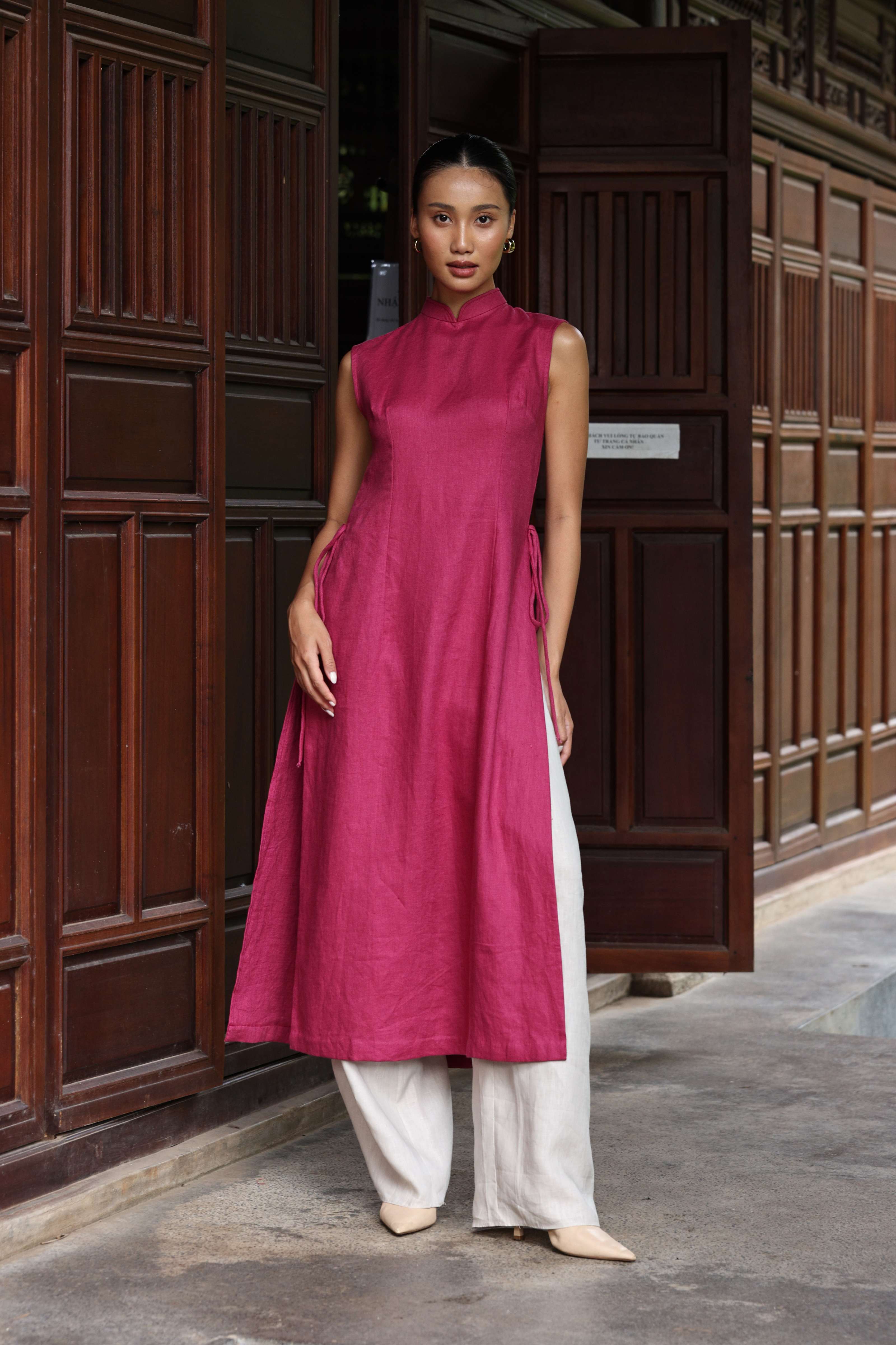 Woman wearing a magenta dress with white pants standing in front of a wooden door.