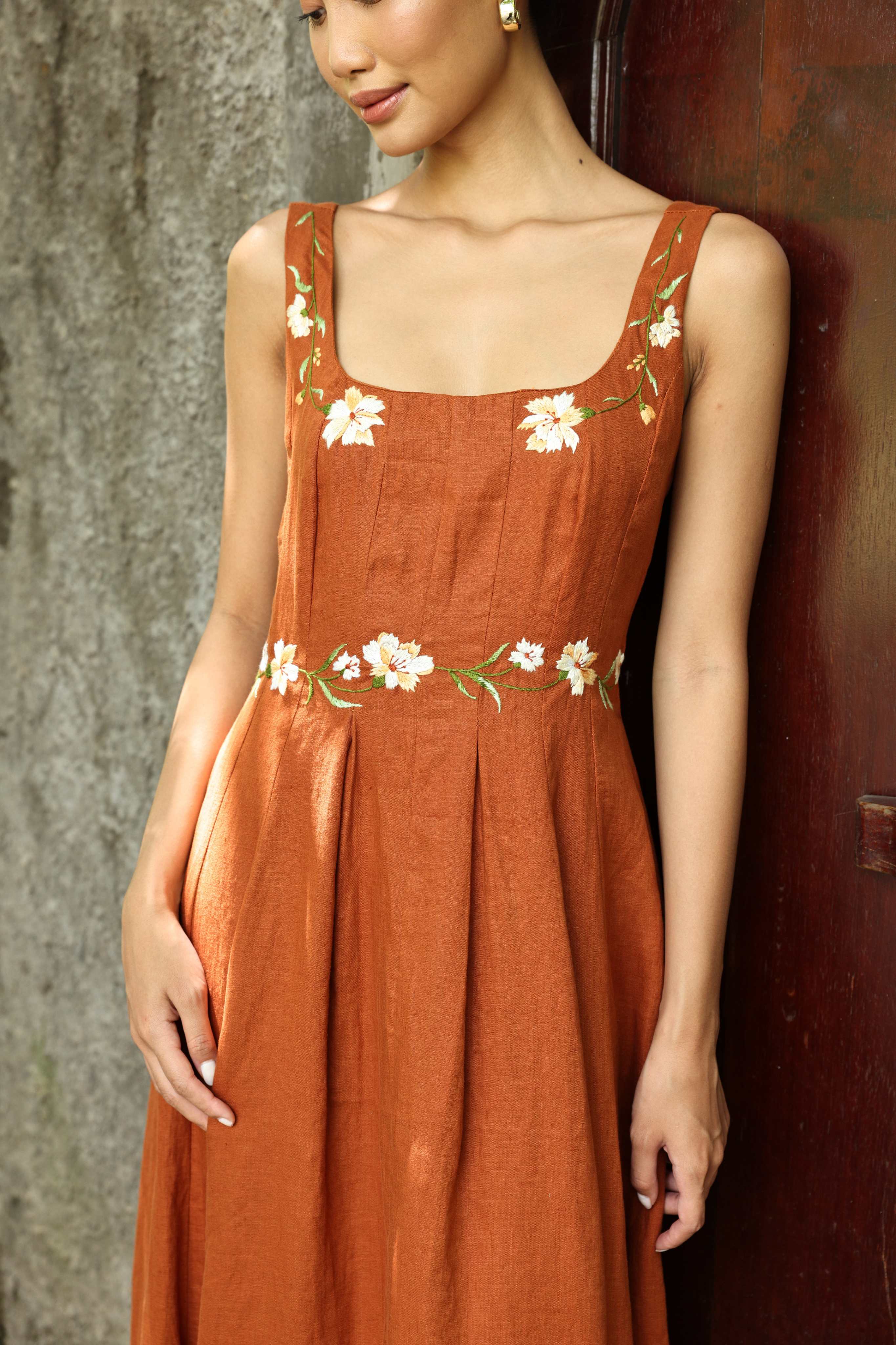 Woman wearing a rust-colored dress with floral embroidery against a textured wall.