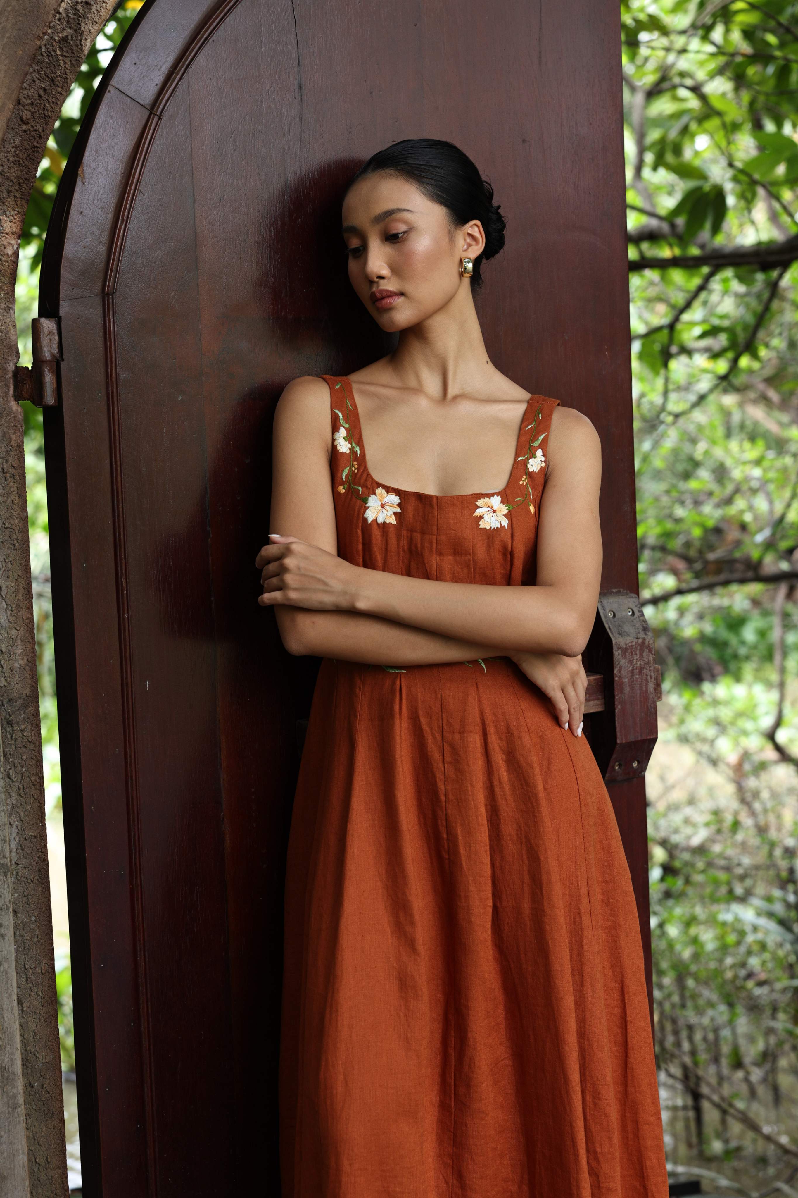 Woman in a rust-colored dress with floral patterns standing against a wooden door with greenery in the background
