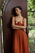 Woman in a rust-colored dress with floral patterns standing against a wooden door with greenery in the background