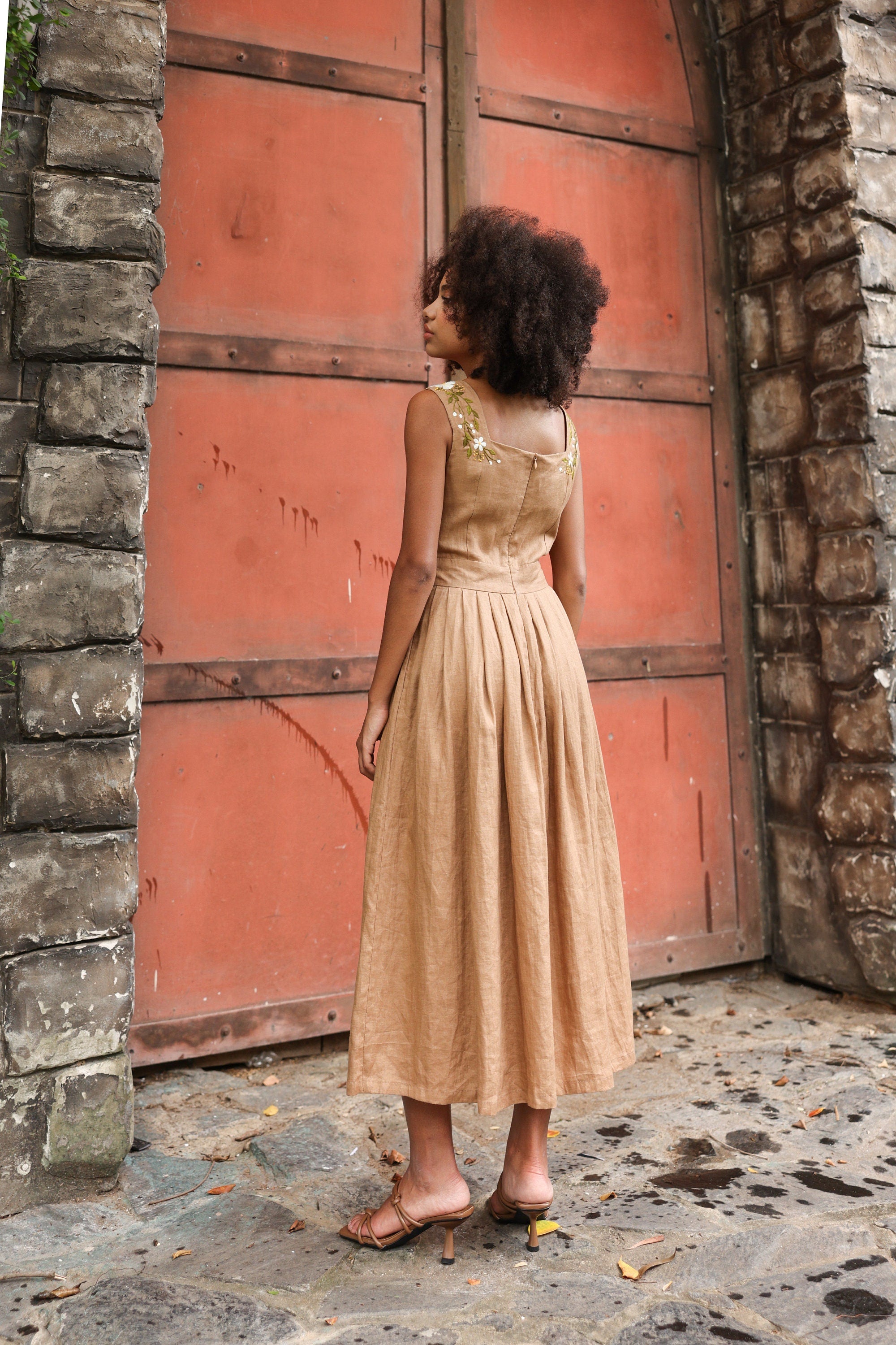 Woman in a dress standing in front of a large red door and stone wall.