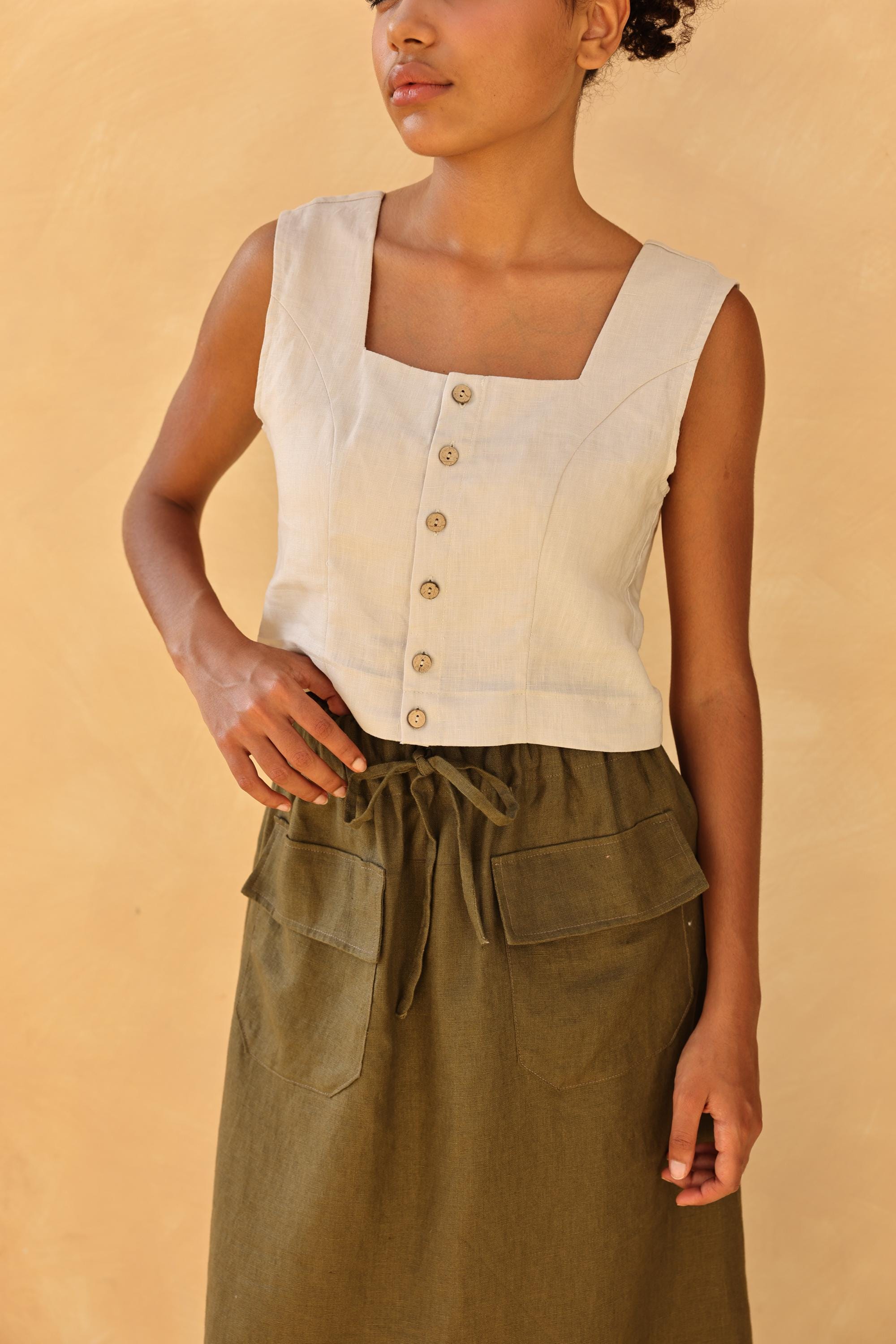 Woman wearing a sleeveless beige top with buttons and a green skirt against a beige background