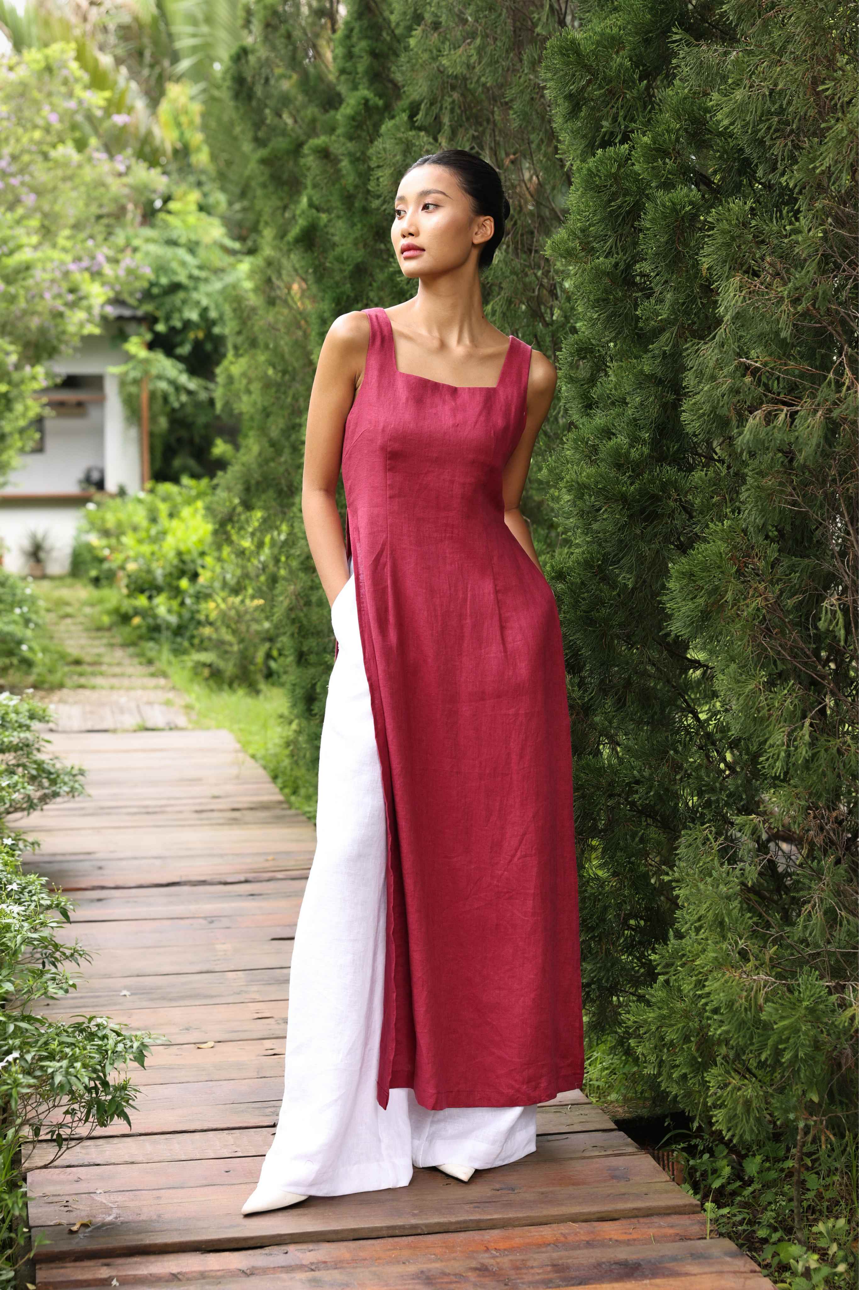 Woman in a red tunic and white pants standing on a wooden path with greenery around.