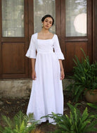 Woman in a white dress standing outdoors with plants and a wooden door in the background
