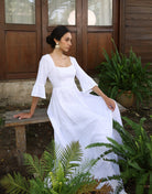 Woman in a white dress sitting on a wooden bench surrounded by greenery.