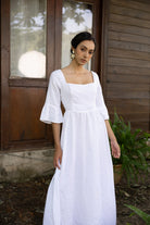 Woman wearing a white dress standing outdoors with wooden walls and plants in the background