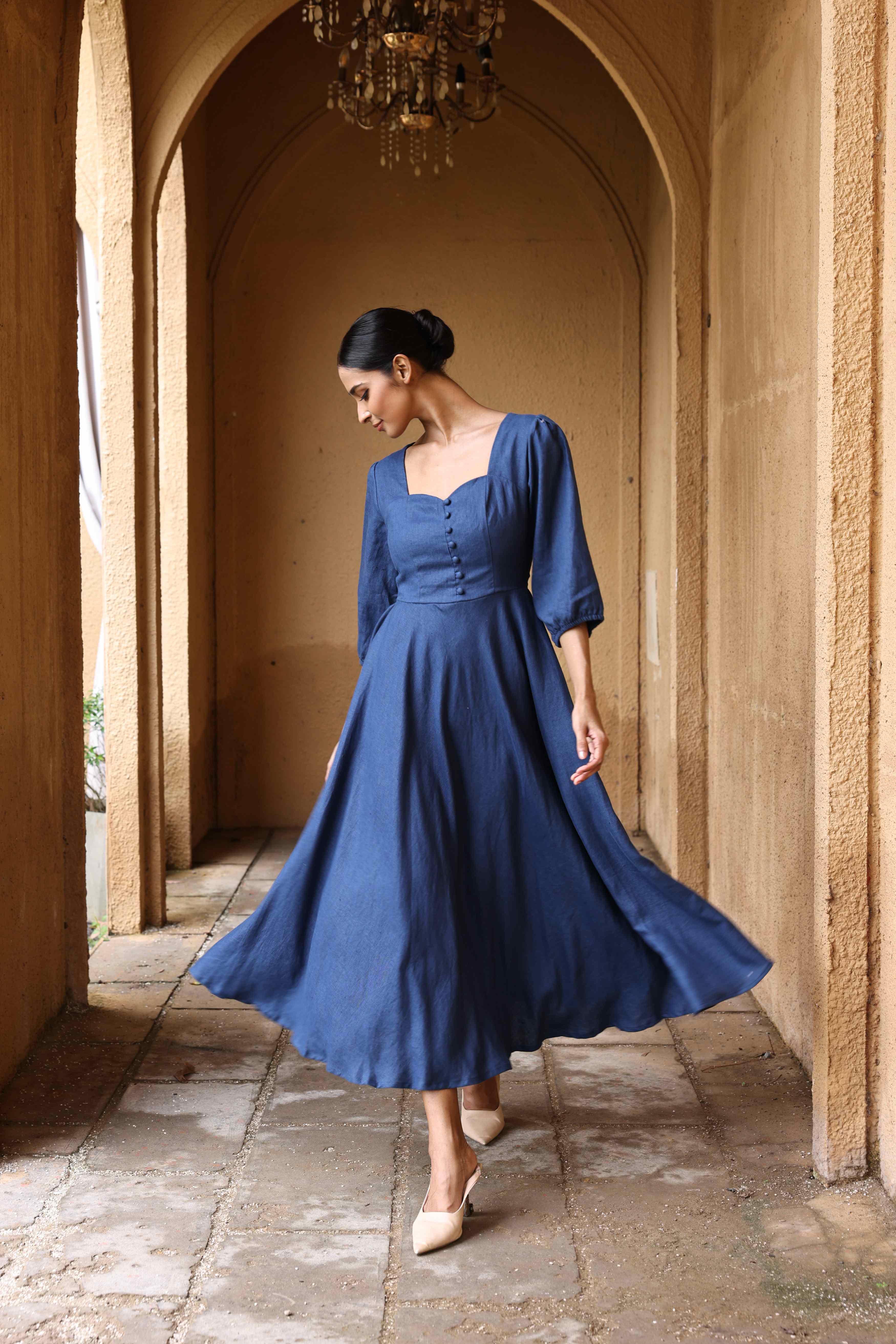 A woman in a blue dress with a high neckline and long sleeves stands in a hallway with arched doorways.