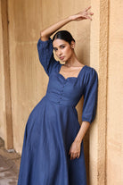 A woman in a blue dress poses with her arm raised against a beige wall.