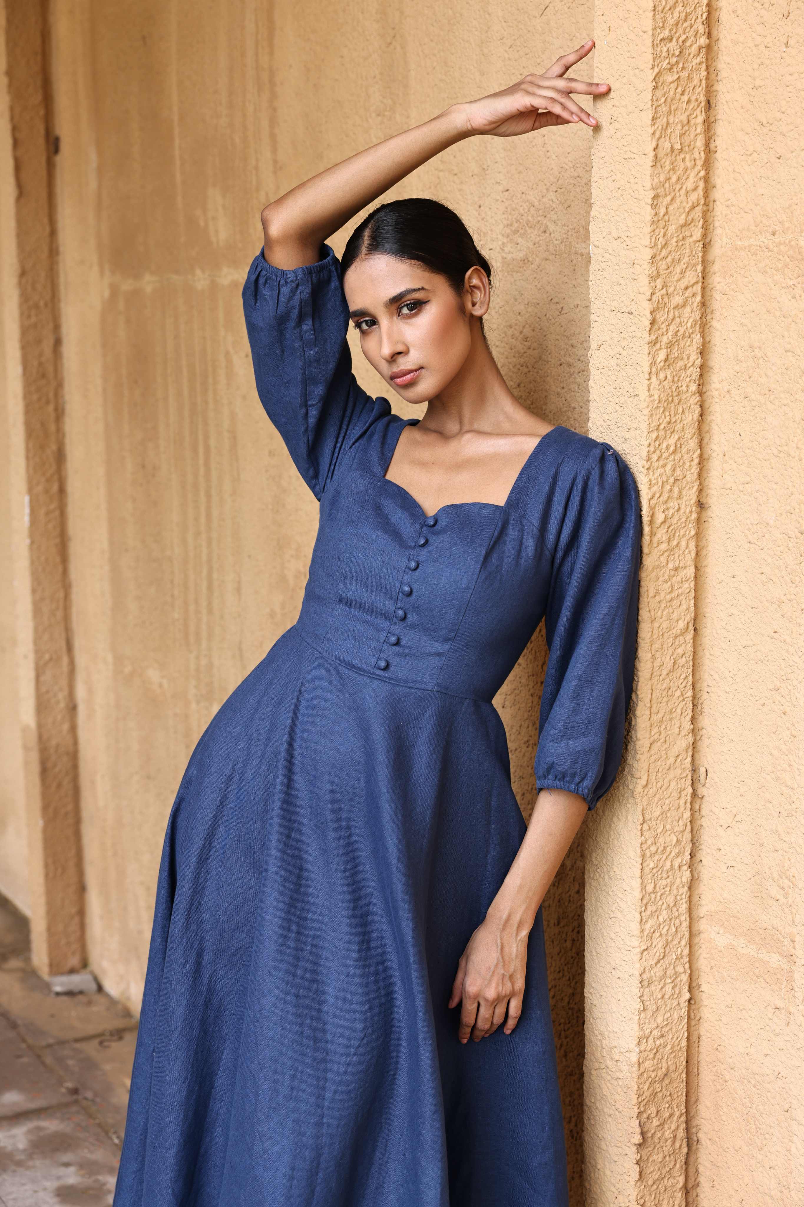 A woman in a blue dress poses with her arm raised against a beige wall.