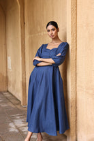 A woman in a blue dress stands with her arms crossed, leaning against a beige wall.