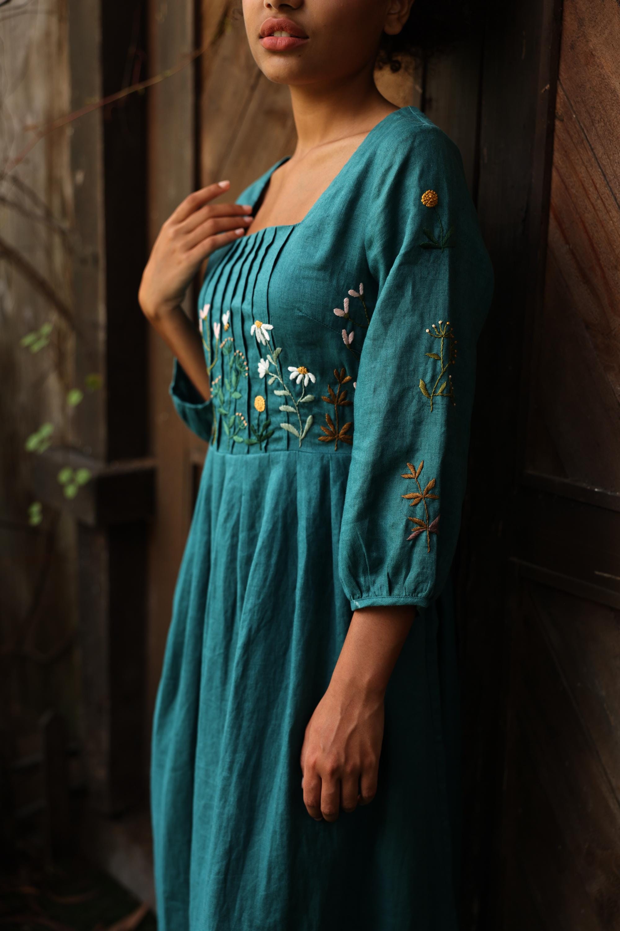 Woman wearing a teal dress with floral embroidery against a wooden wall.