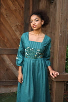 Woman wearing a teal dress with floral embroidery against a wooden background