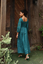 Woman in a teal dress standing in front of a wooden door with plants around.