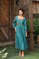 Woman in a teal dress standing outdoors with wooden walls and greenery in the background