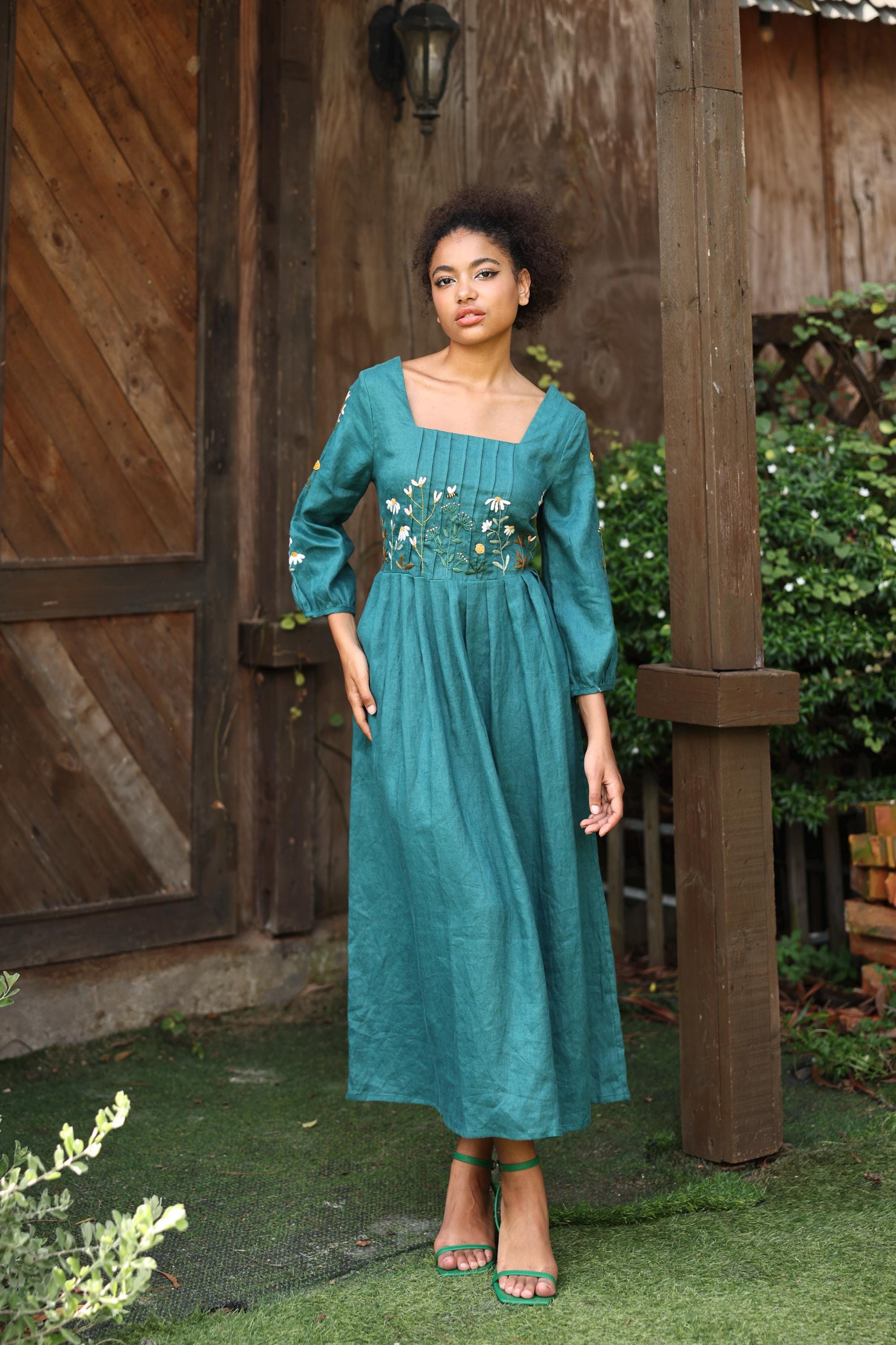 Woman in a teal dress standing outdoors with wooden walls and greenery in the background