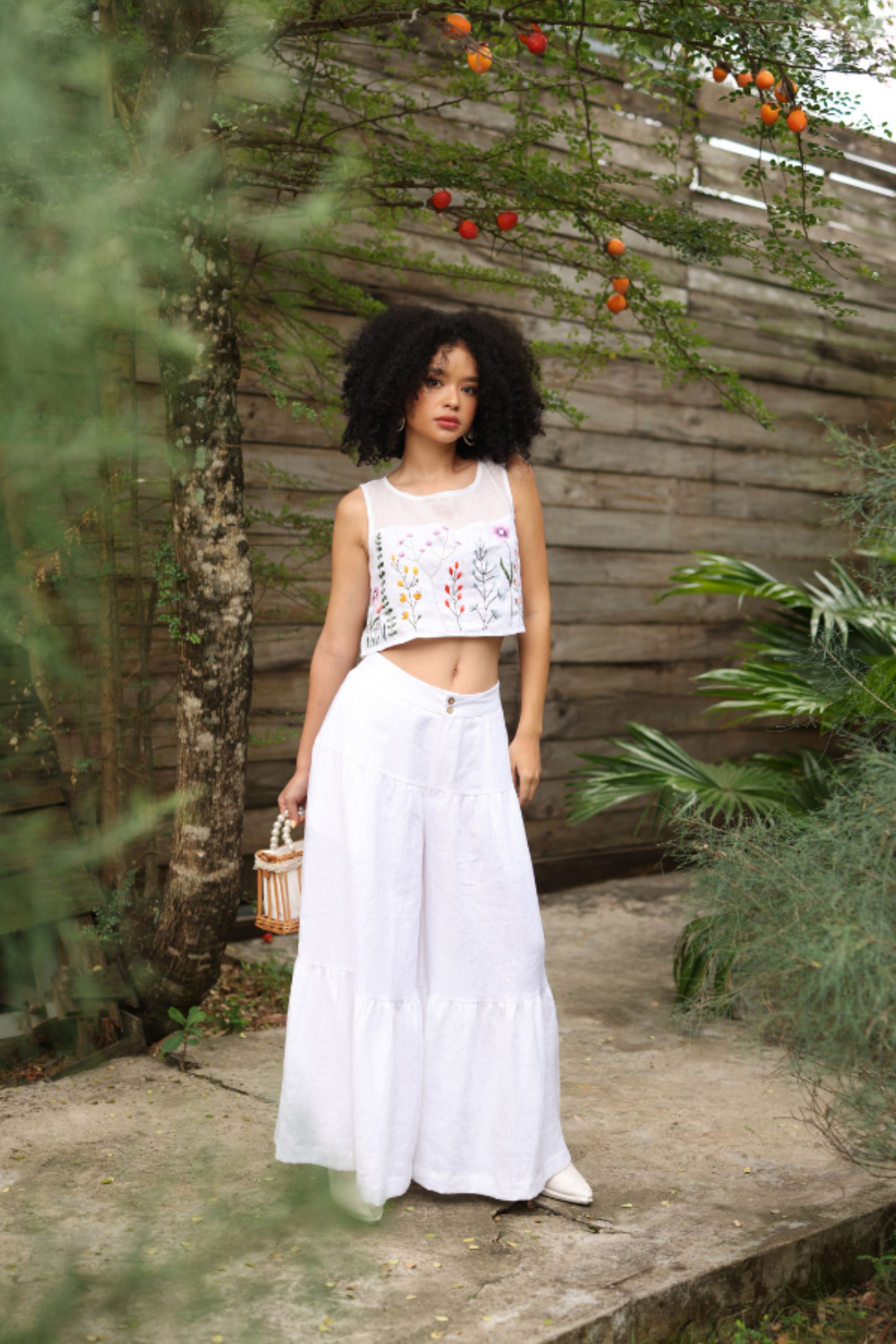 Woman in a white outfit with floral top and long white pants standing in a garden.