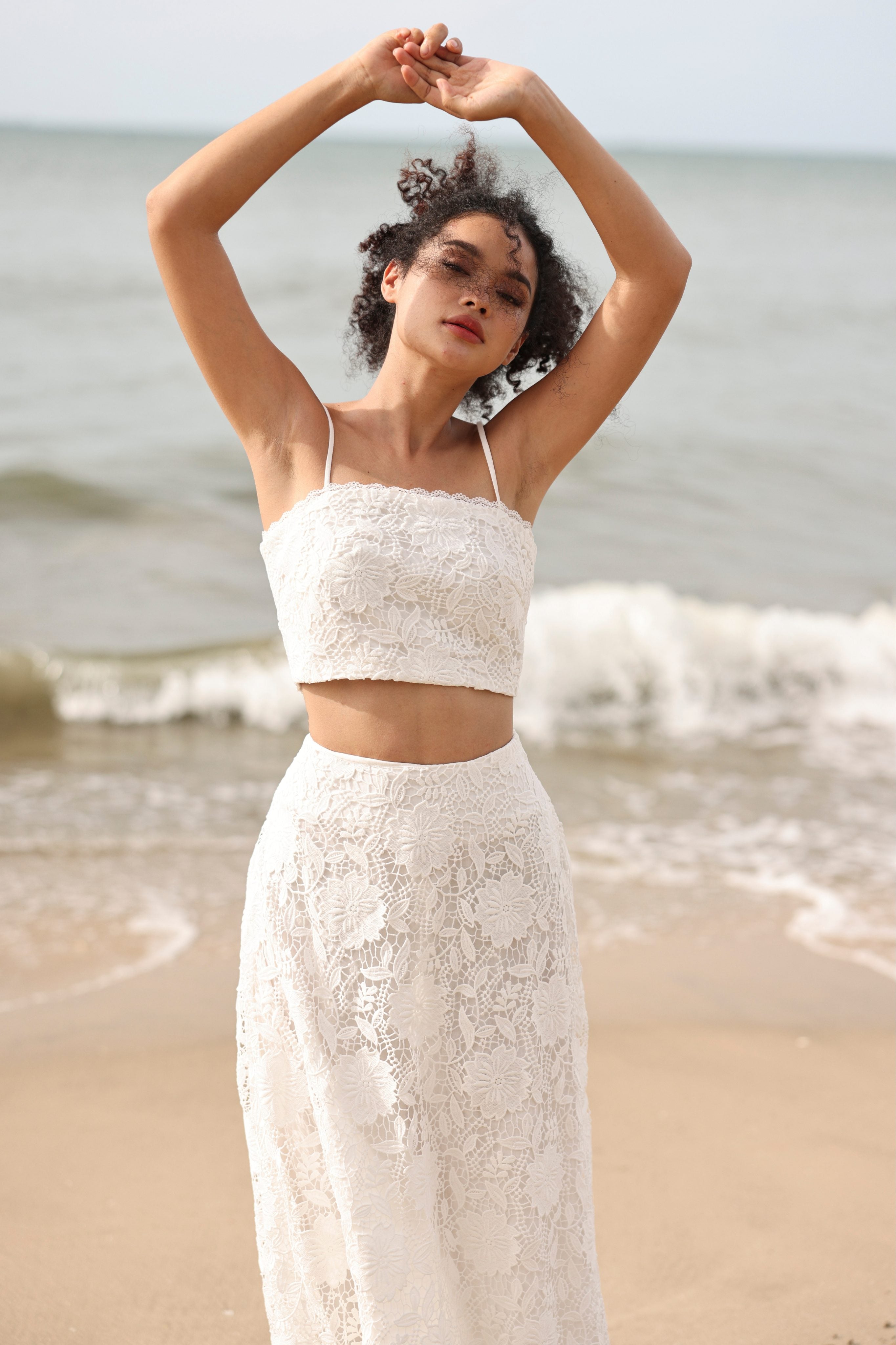Woman in a white lace outfit standing on a beach with ocean waves in the background