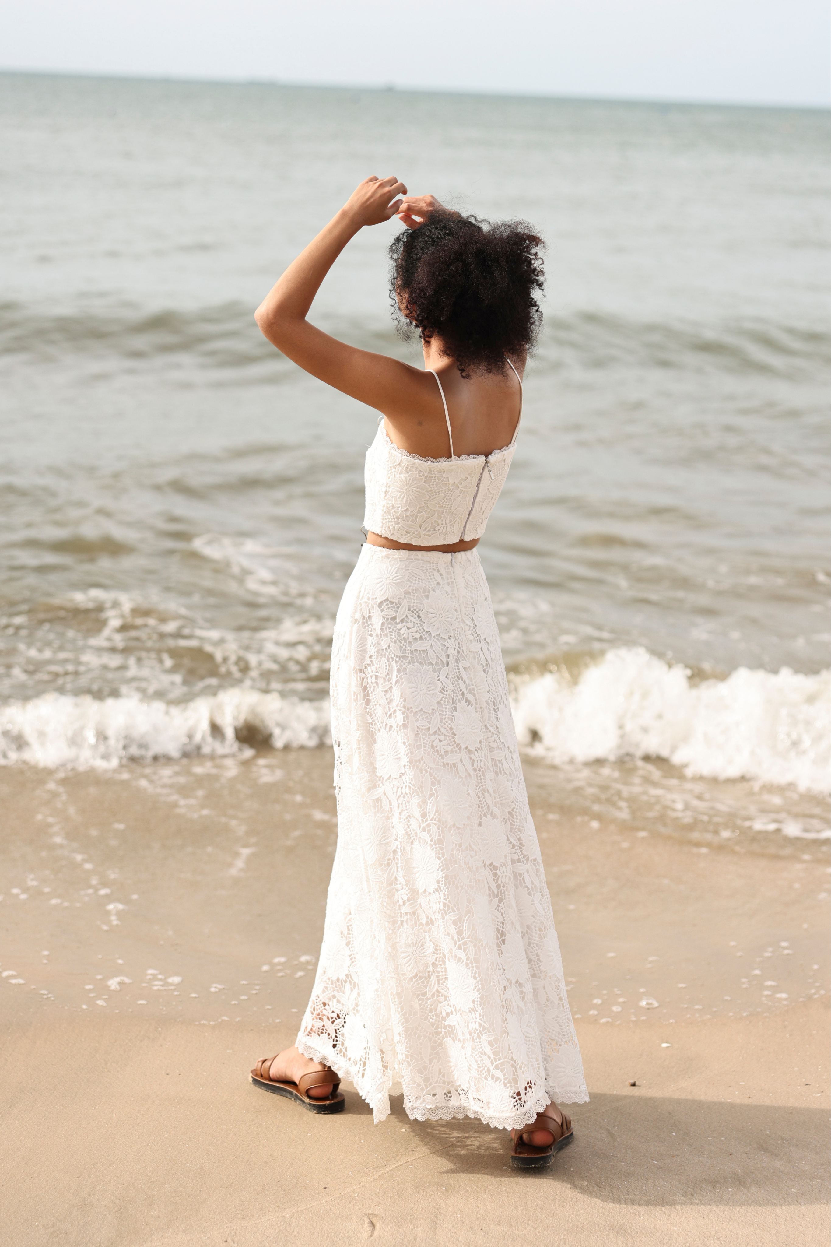 Woman in a white lace top and skirt standing on a beach with ocean waves.