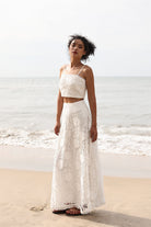 Woman in a white lace top and skirt standing on a beach with ocean in the background