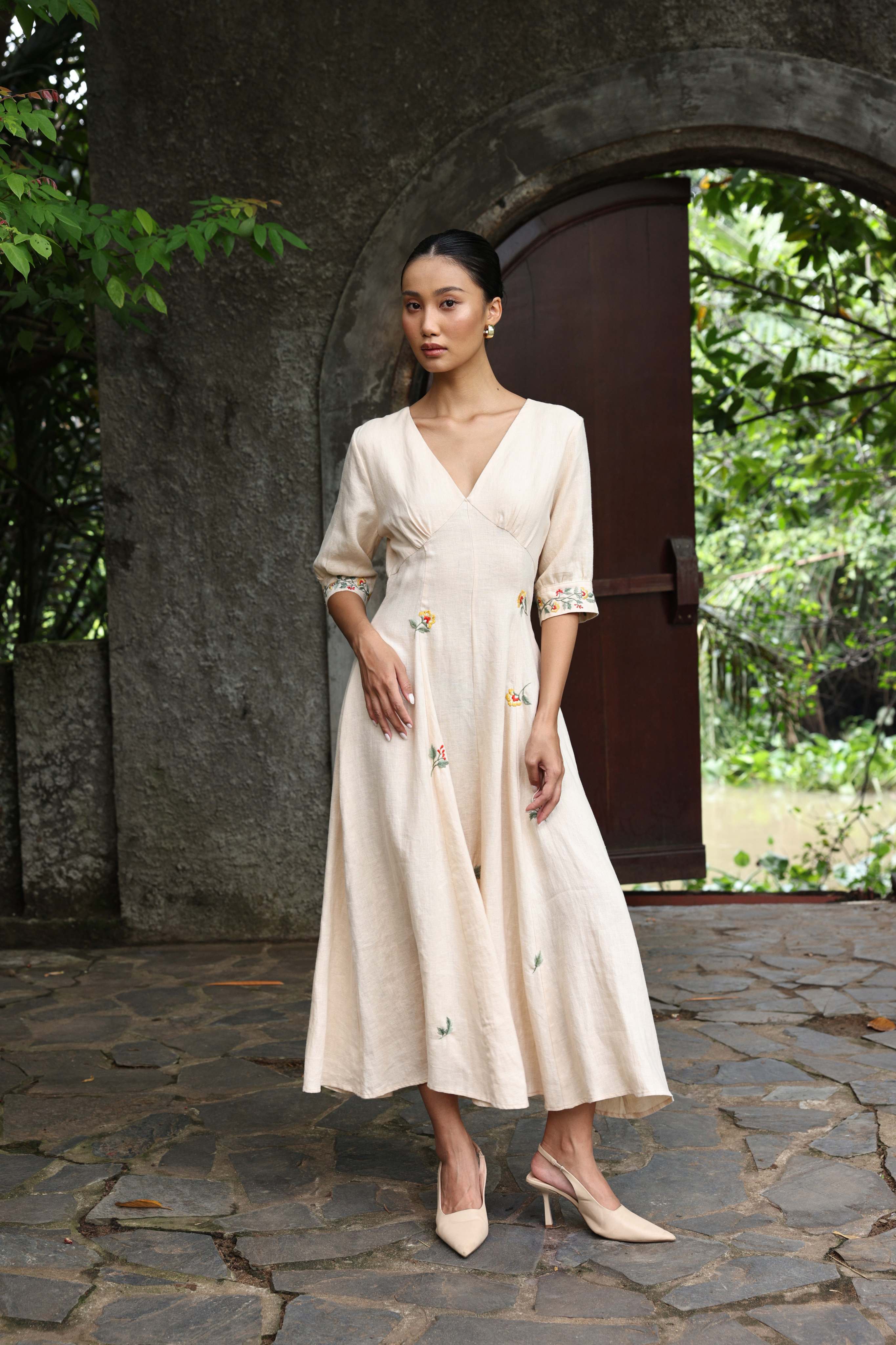 Woman in a beige dress standing in front of a stone archway with greenery.