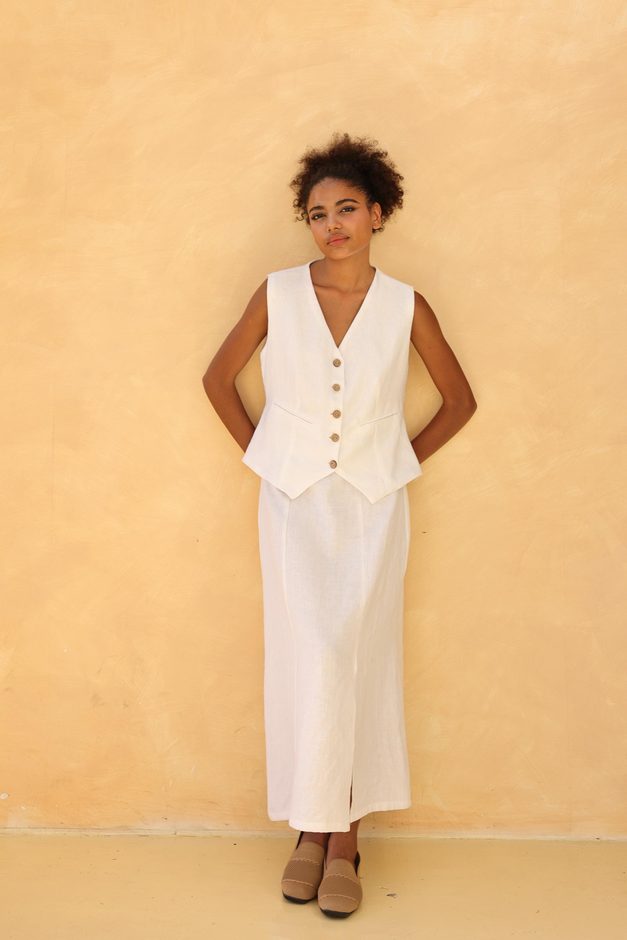 Woman wearing a white sleeveless vest over a long white skirt against a beige wall.